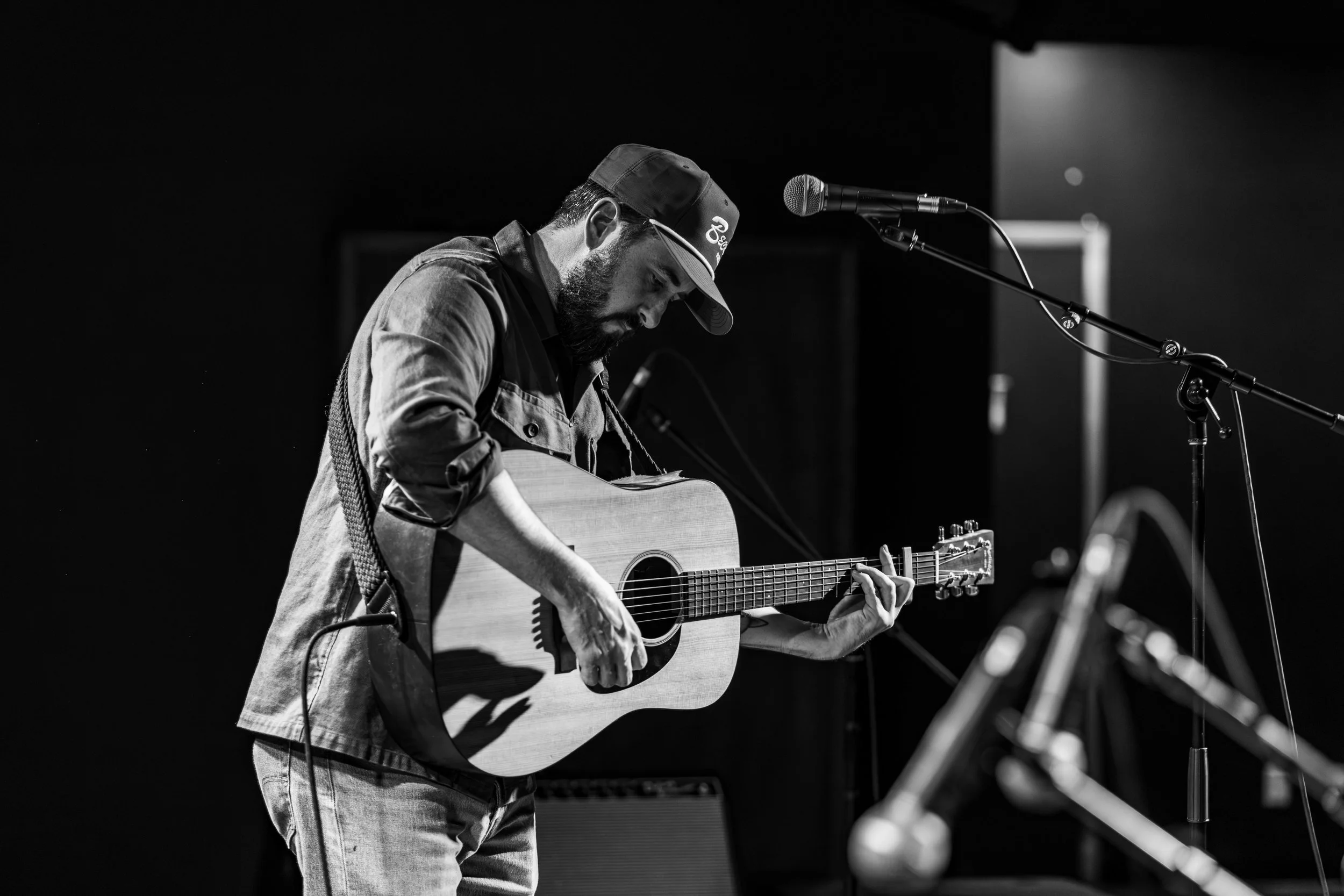 Black and white photo of a man playing an acoustic guitar on stage, with a microphone nearby, in a dimly lit setting.