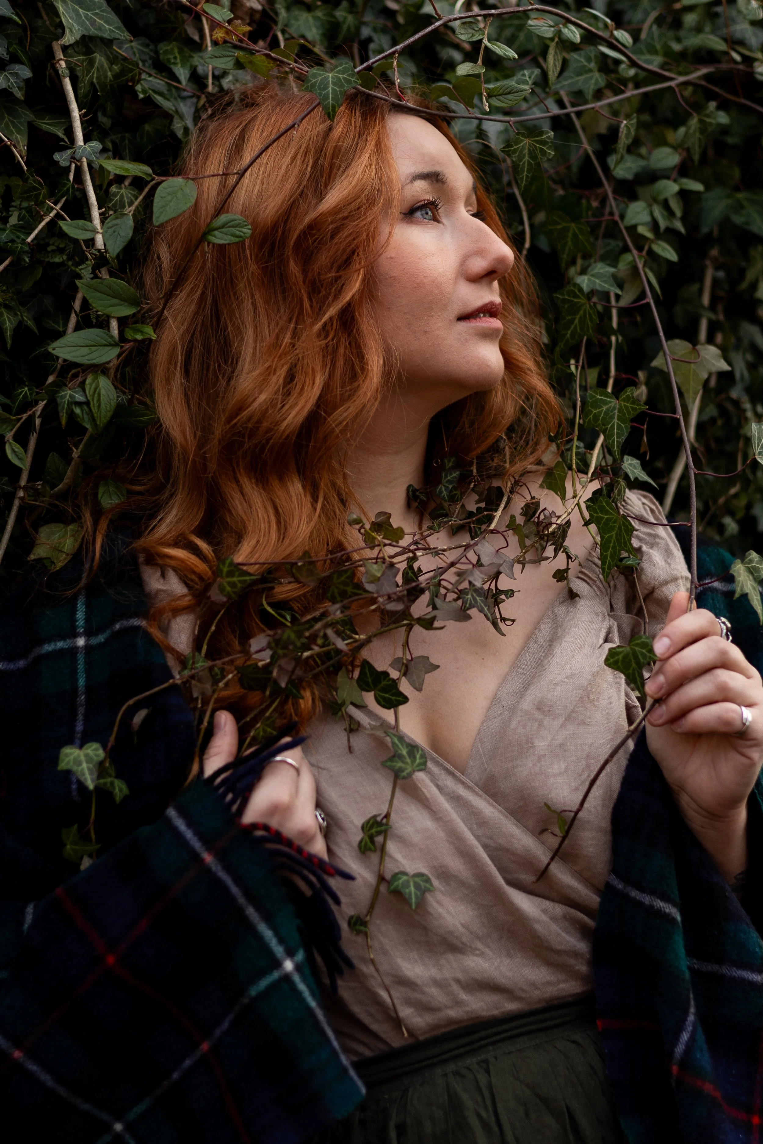 A woman with long, wavy red hair looking to the side among dense green ivy leaves.