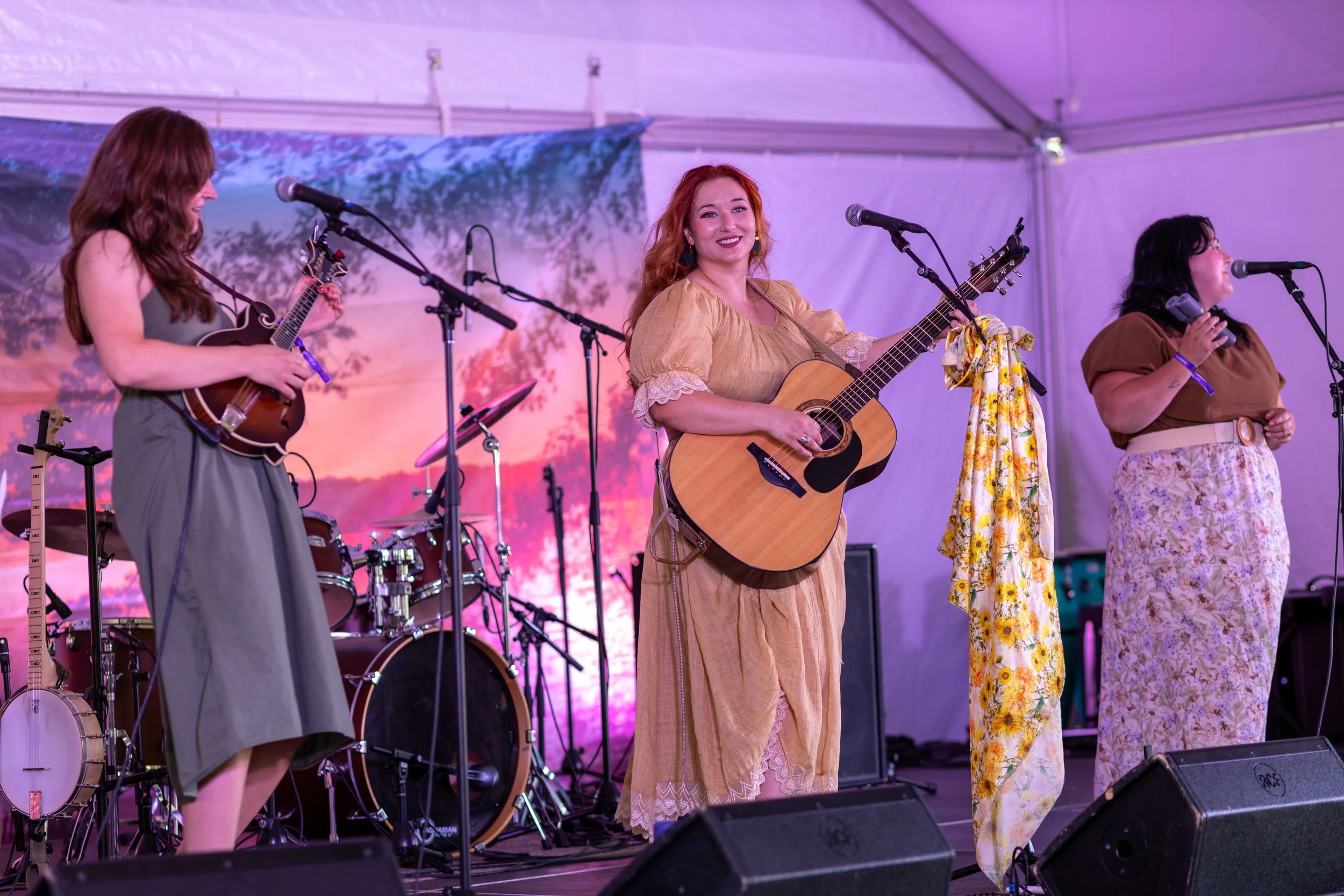 Four women performing on stage with musical instruments, including guitars and drums, in a tent with a scenic backdrop and colorful lighting.