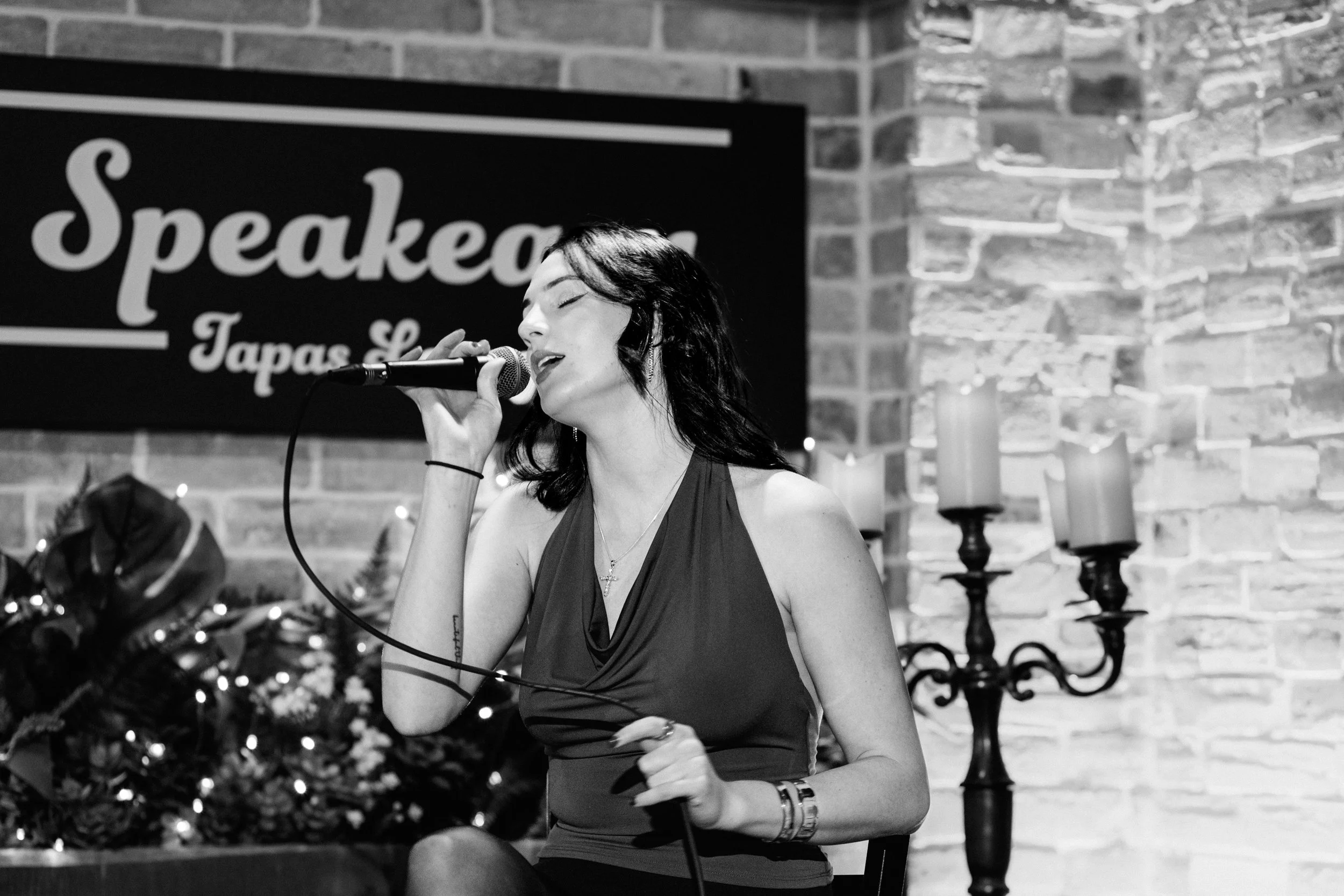 A woman singing into a microphone during a performance at a jazz lounge, with a sign reading 'Speakeasy Tapas Lounge' in the background and candles on a candle holder.