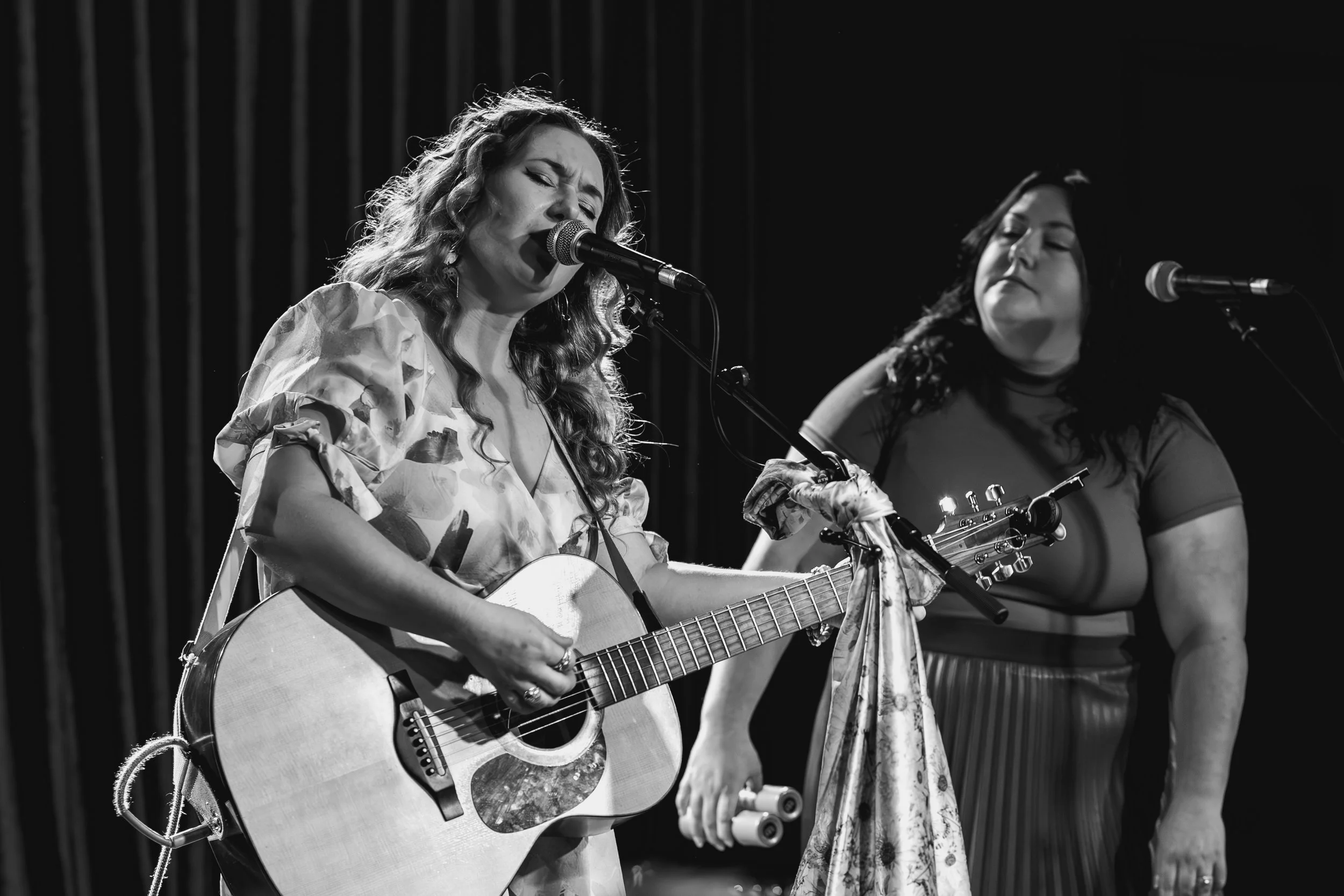 Two women performing on stage; one is singing into a microphone and playing an acoustic guitar, the other is standing nearby with her eyes closed.