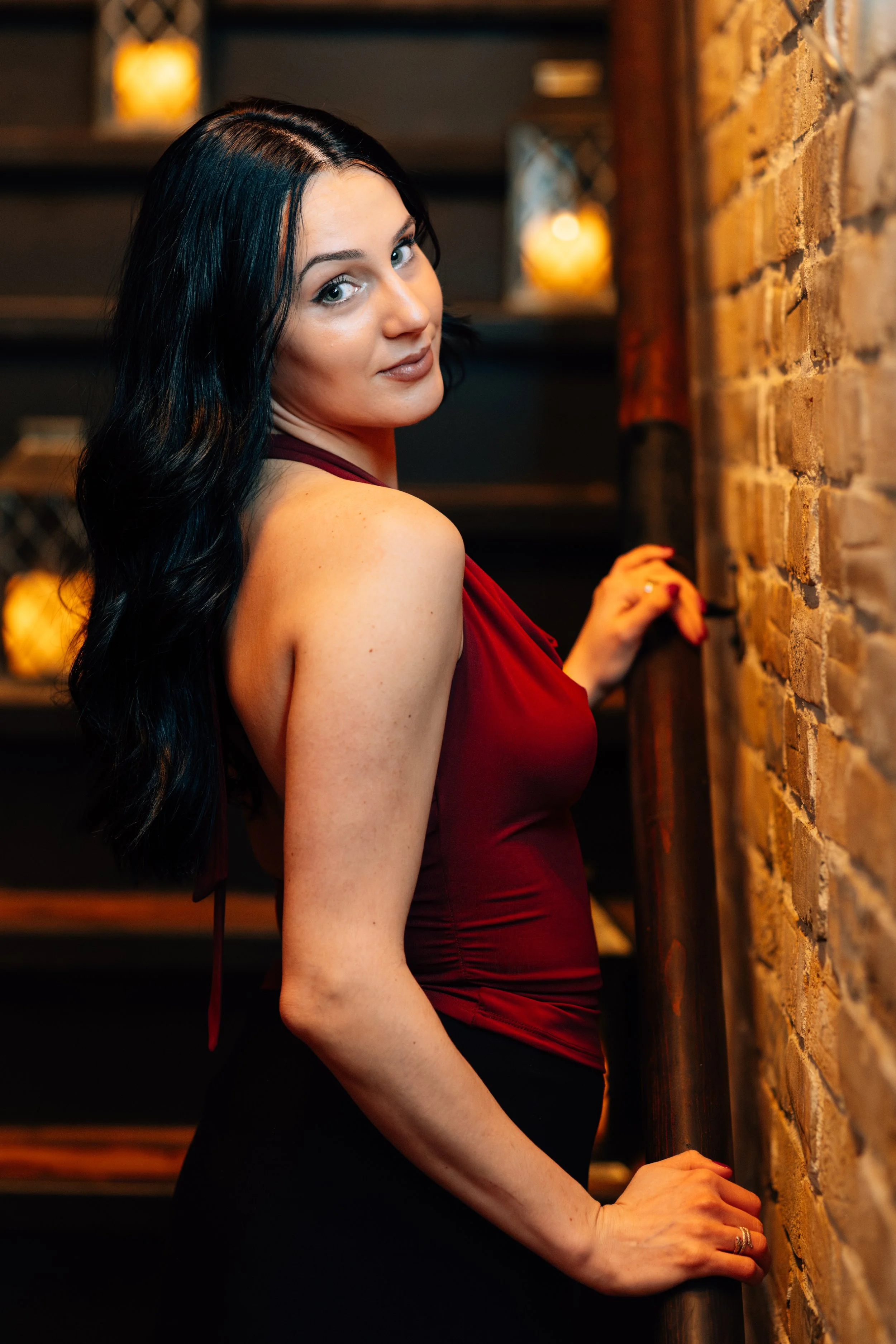 A woman with long black hair and fair skin, wearing a red sleeveless top, standing indoors near a brick wall with warm lighting in the background.