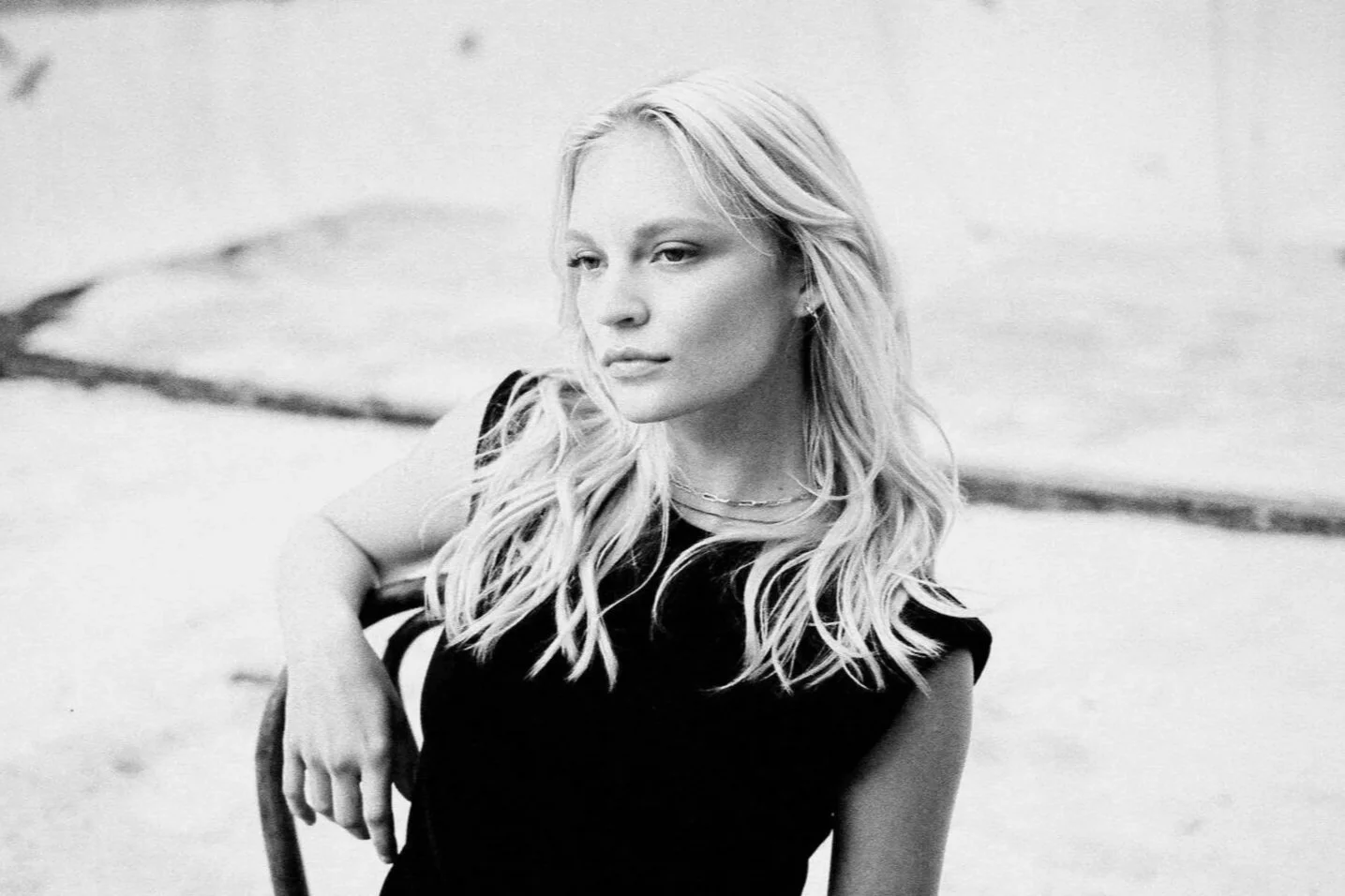 Black and white photo of a woman with long, wavy blonde hair, wearing a sleeveless black top, sitting on a chair outdoors.