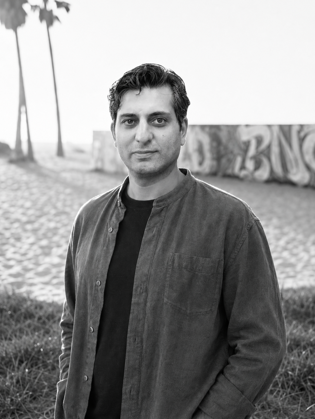 A man with dark hair wearing a brown shirt and black t-shirt standing outdoors with buildings and trees in the background.