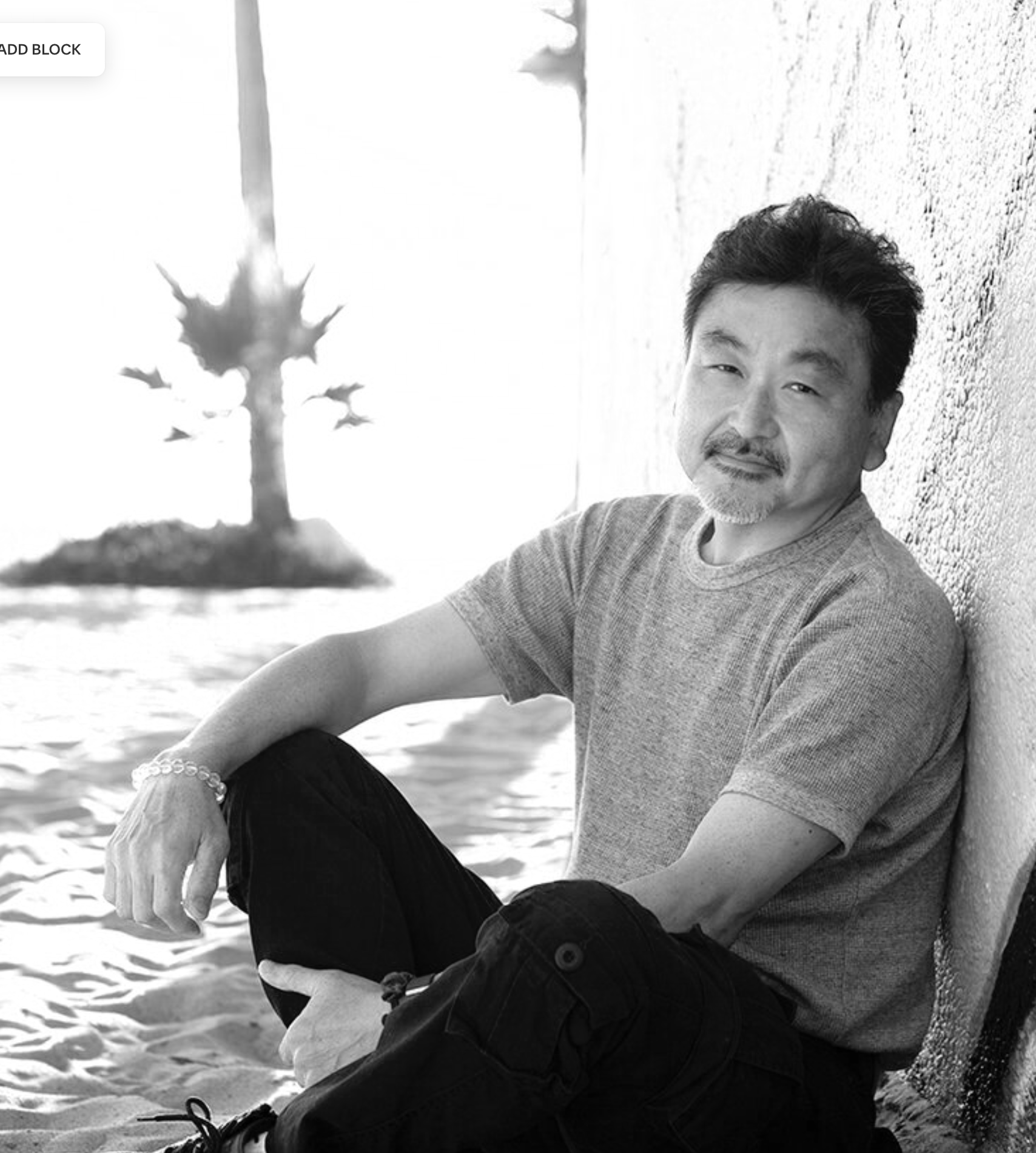 A middle-aged Asian man sitting on the sandy beach near a colorful wall, with a palm tree in the background, during sunset.