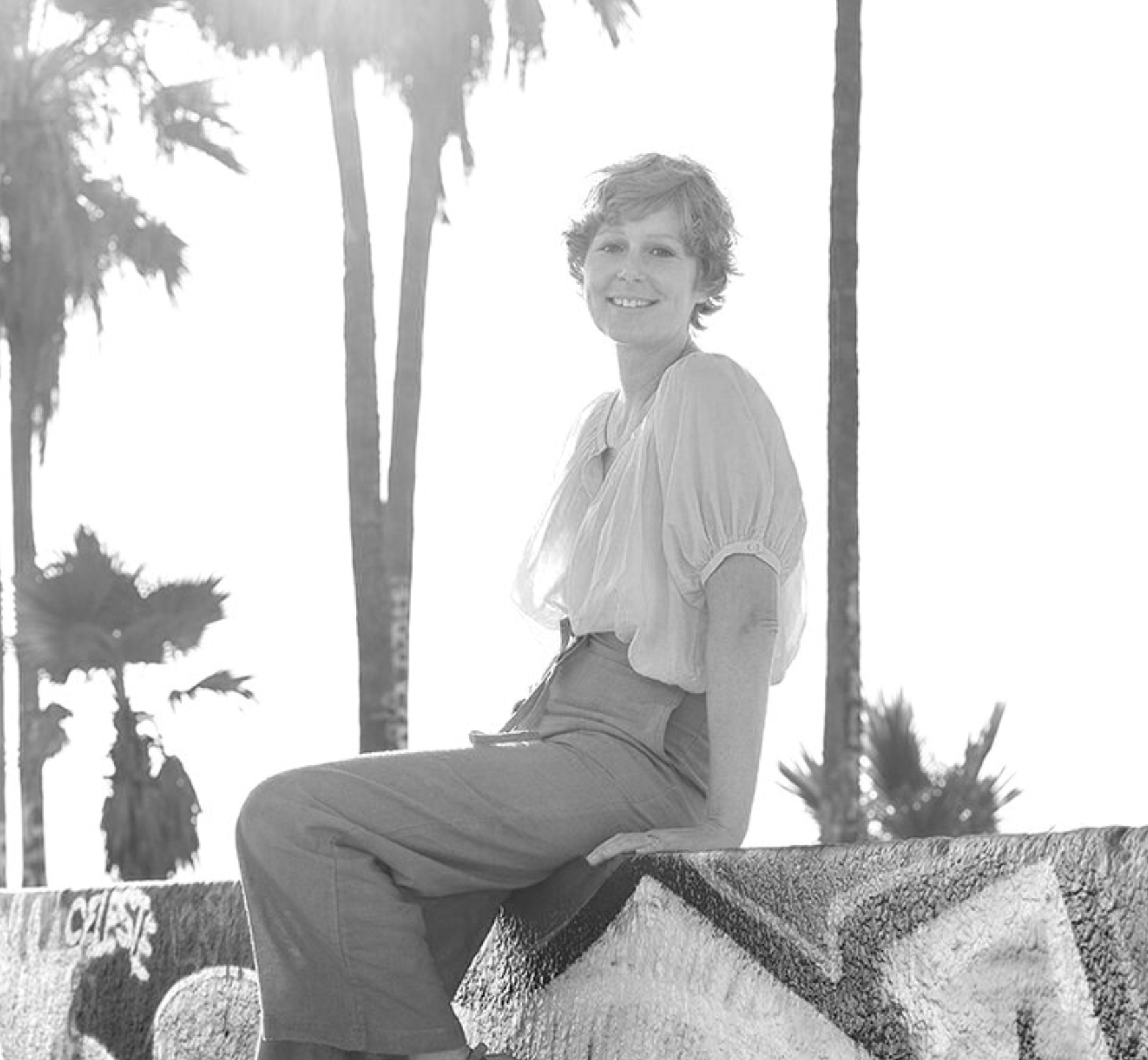 A woman with short wavy hair sitting on a colorful mosaic wall outdoors, smiling, with tall palm trees in the background during sunset.