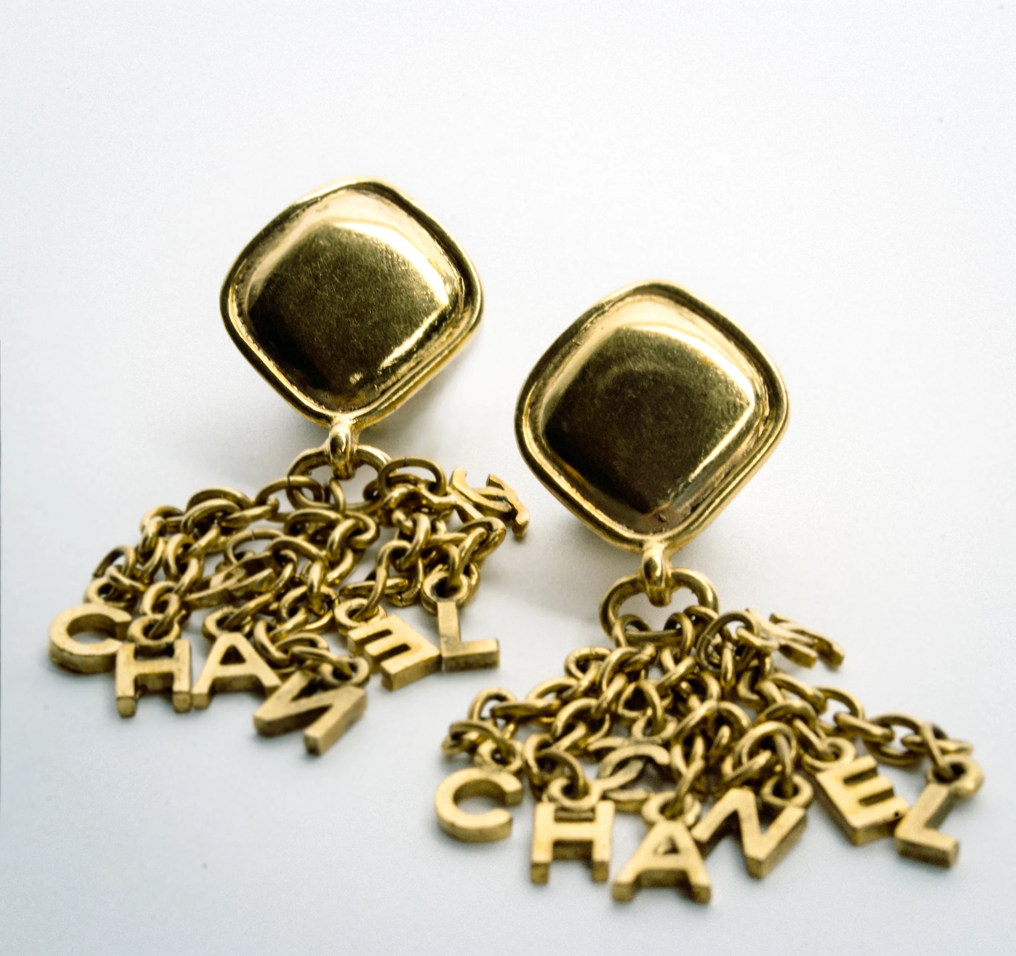 Gold earrings with chain and Chanel letters design on a plain background.