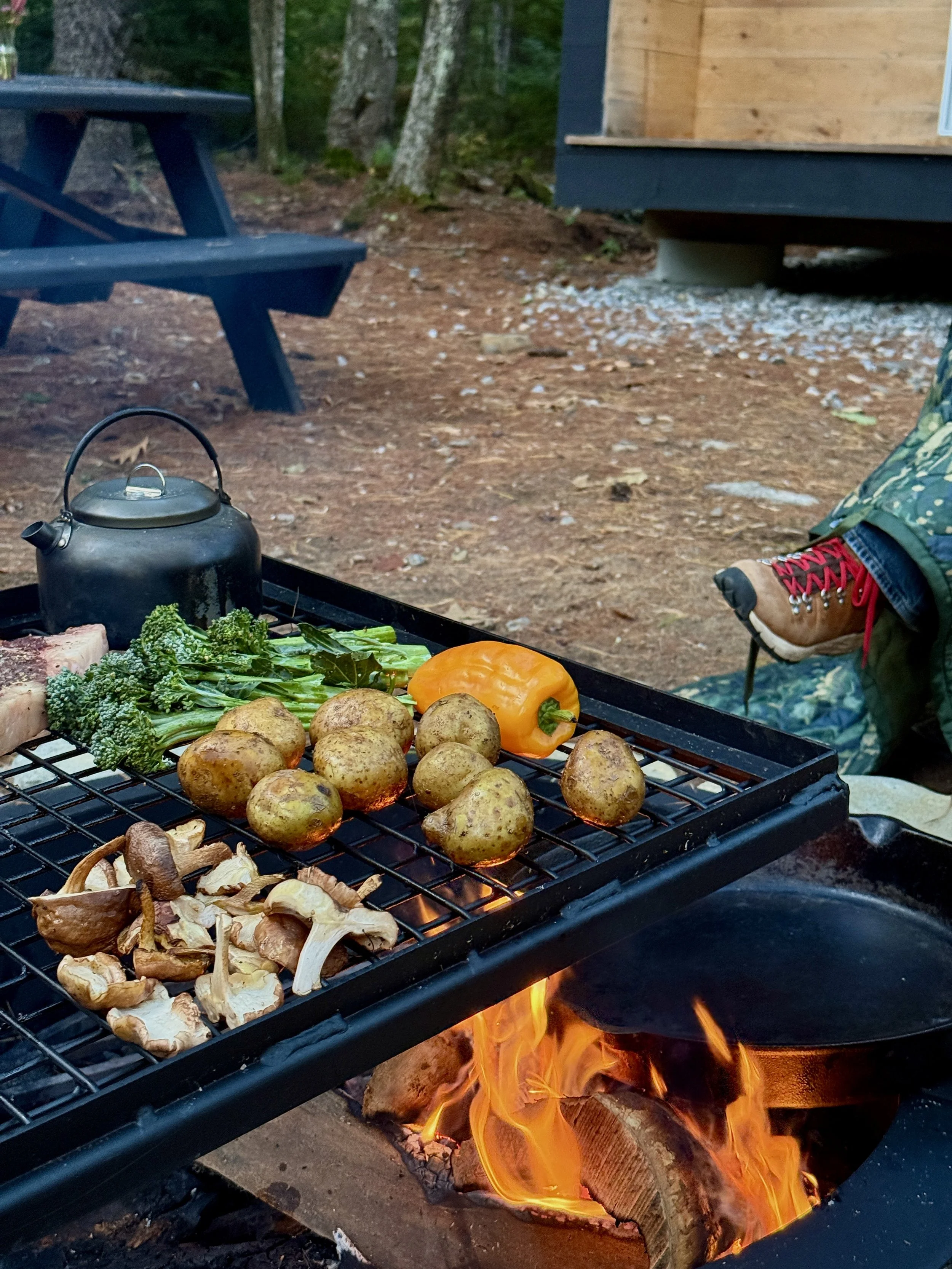 Applecroft Cabin in background with open pit and flame cooking mushrooms, broccolini, bell pepper, potatoes, and meat with kettle and hiking boot in frame