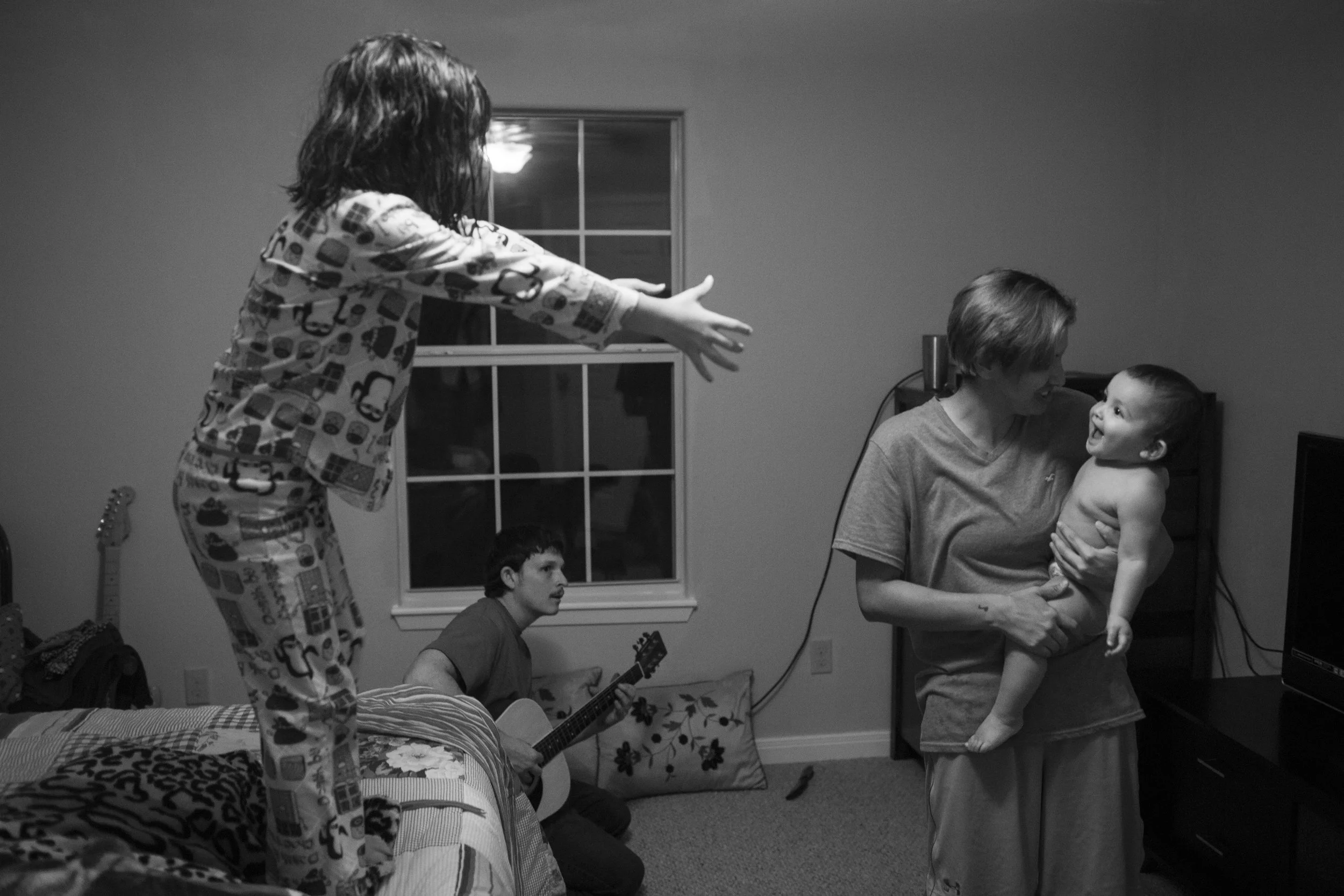  While Jarrell Bamberger plays guitar, Serenity Bamberger stands on her parents' bed and reaches out towards her sister Esme Bella Bamberger, who is in Glenda Bamberger's arms, on their first official day living in their new home in Blanco, Texas on 