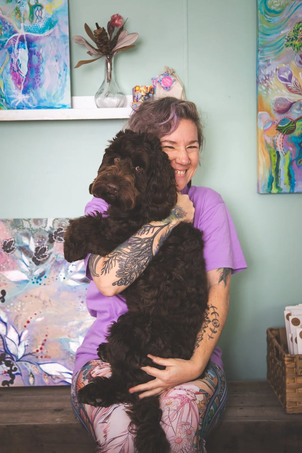 A woman with tattoos and purple hair holding a brown curly-haired puppy in an art studio with colorful paintings and a plant on a shelf in the background.