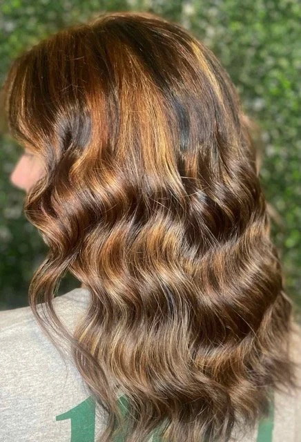 Back view of long, wavy brunette hair with warm copper and caramel highlights. Styled at a Seattle hair salon for rich dimension and healthy shine.
