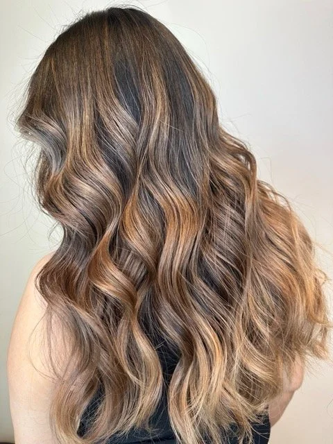 Long brunette balayage with warm caramel highlights and soft waves, styled at Chroma Salon for natural dimension and shine.