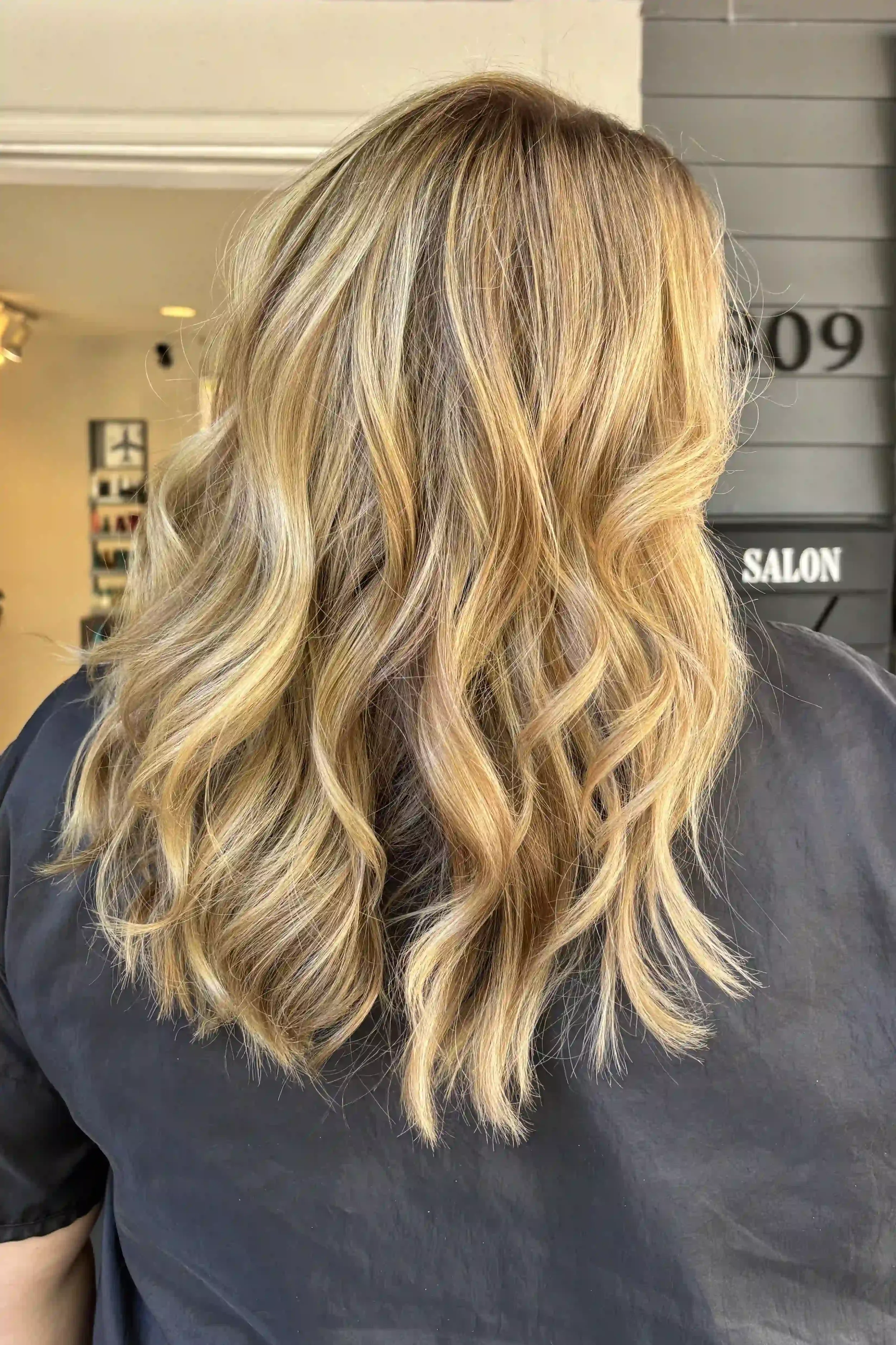 Dimensional golden blonde hair with bright, buttery highlights and warm honey tones that blend into a deeper, natural-looking base. Medium-length layers and loose, tousled waves create soft movement, texture, and fullness. Hair by Alpin at Chroma Sal