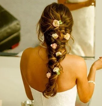 Romantic bridal hairstyle featuring a softly textured braid adorned with delicate floral accents. This elegant wedding updo combines loose volume, face-framing softness, and intricate detail for a timeless, romantic look perfect for bridal celebratio