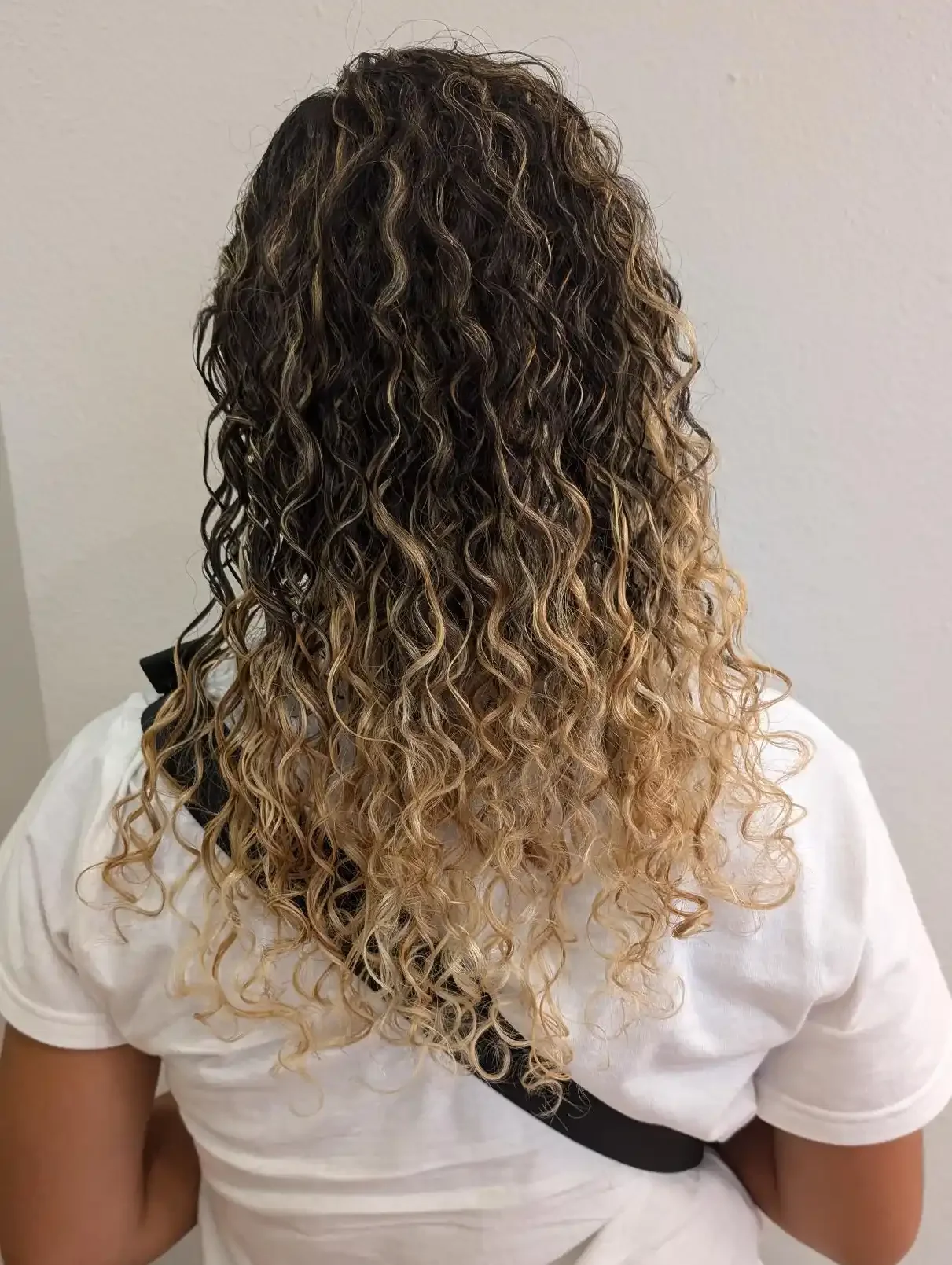 Dimensional blonde balayage on naturally curly hair with bright ribbon highlights and a soft lived-in root melt. Defined spiral curls showcase contrast, depth, and texture while maintaining a natural, low-maintenance finish.