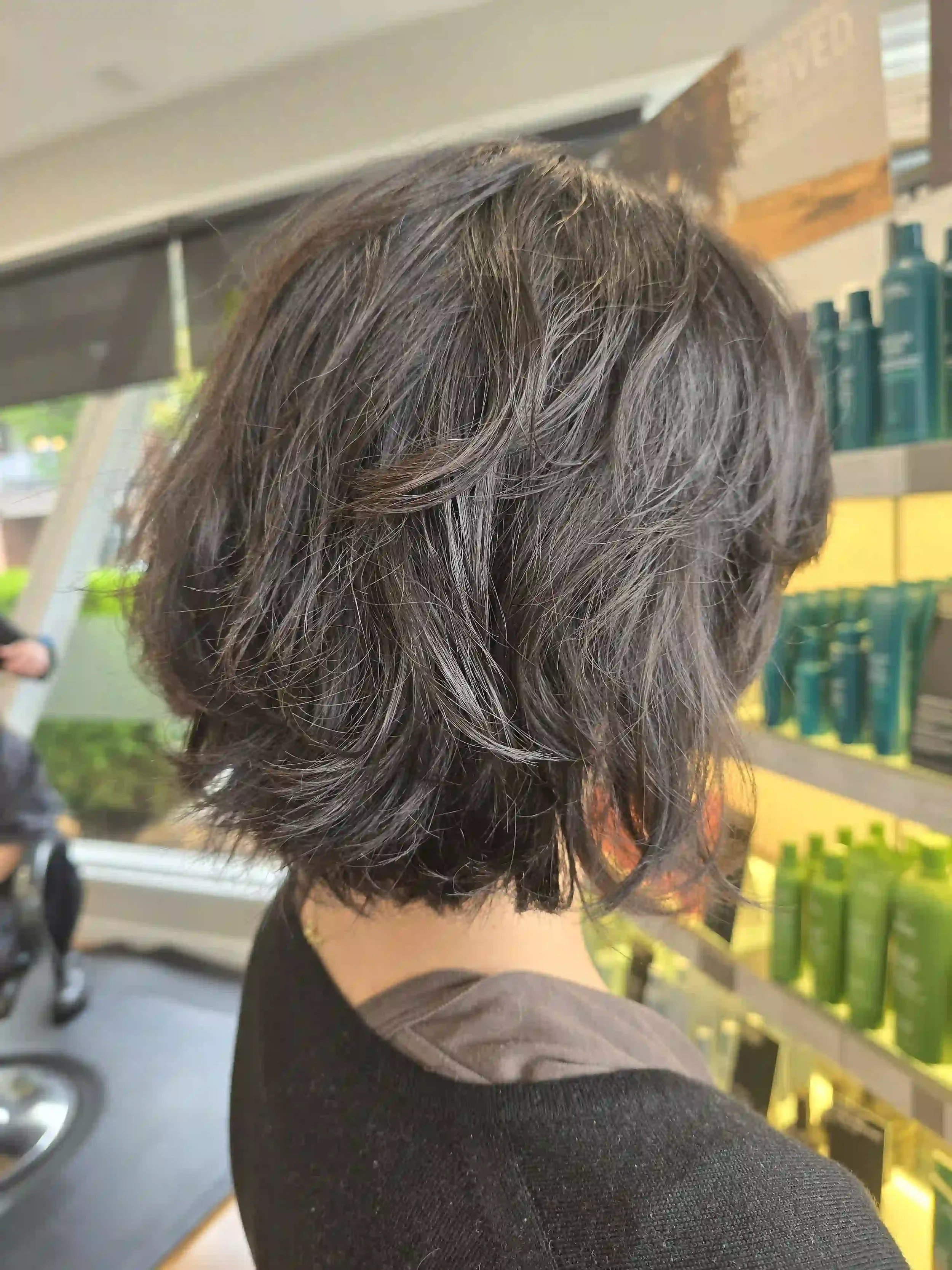 Rich, dark brunette hair in a textured, chin-length bob with soft, choppy layers and feathered ends. The tousled styling creates airy volume and natural movement, giving this layered haircut a relaxed, effortlessly modern finish. Hair by Jhana at Chr