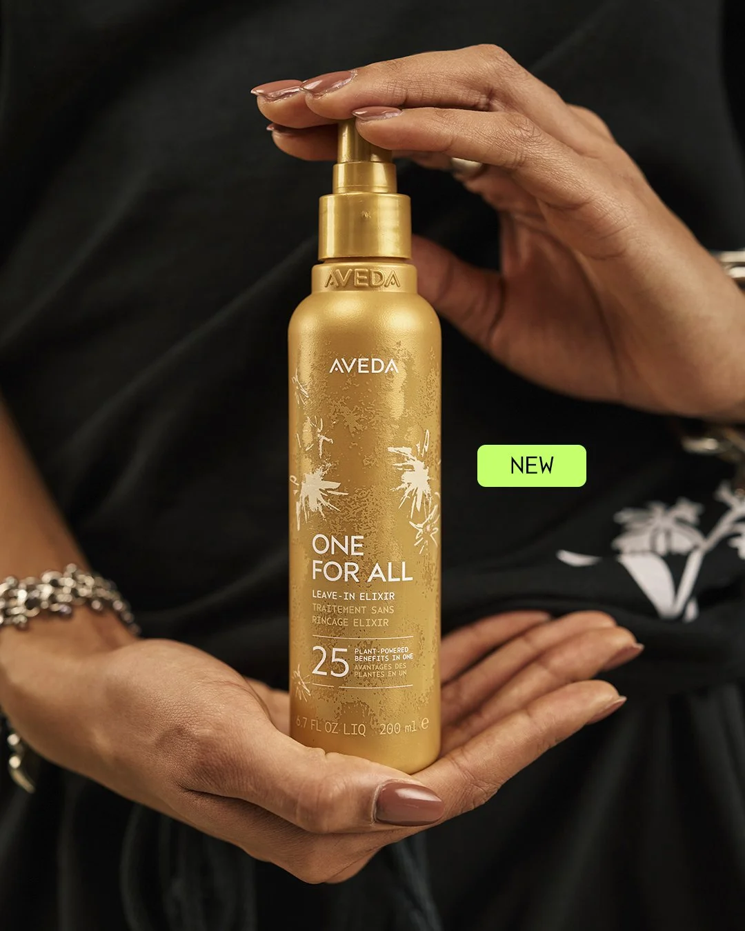 Two hands present a gold bottle of Aveda One For All Leave-In Elixir against a black background, with a bright green “NEW” label beside it.