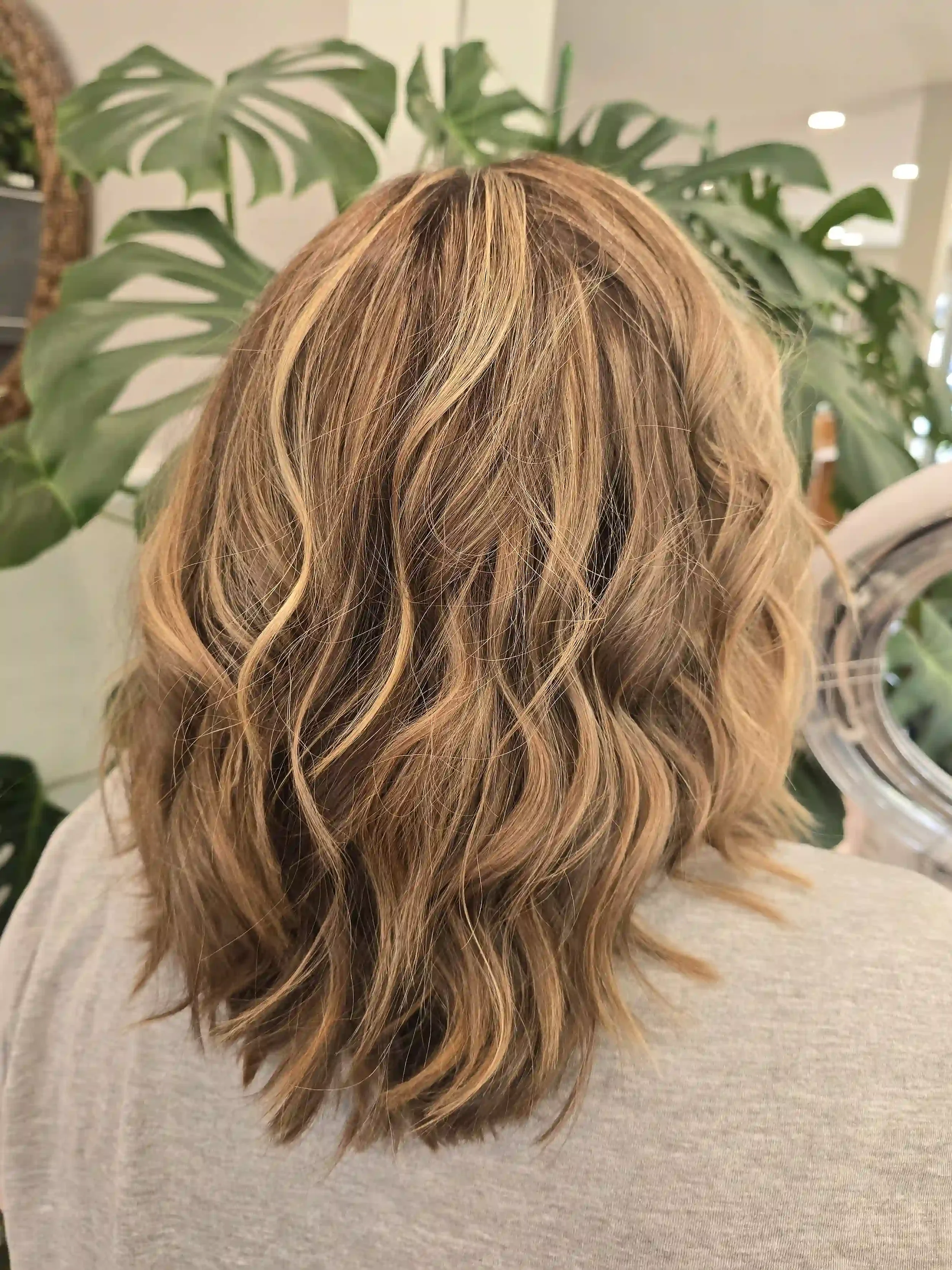 Medium-length bronde balayage with dimensional honey blonde highlights, a soft shadow root, and textured layers styled in effortless beach waves. This lived-in color creates natural depth, brightness, and movement for a low-maintenance, modern look. 