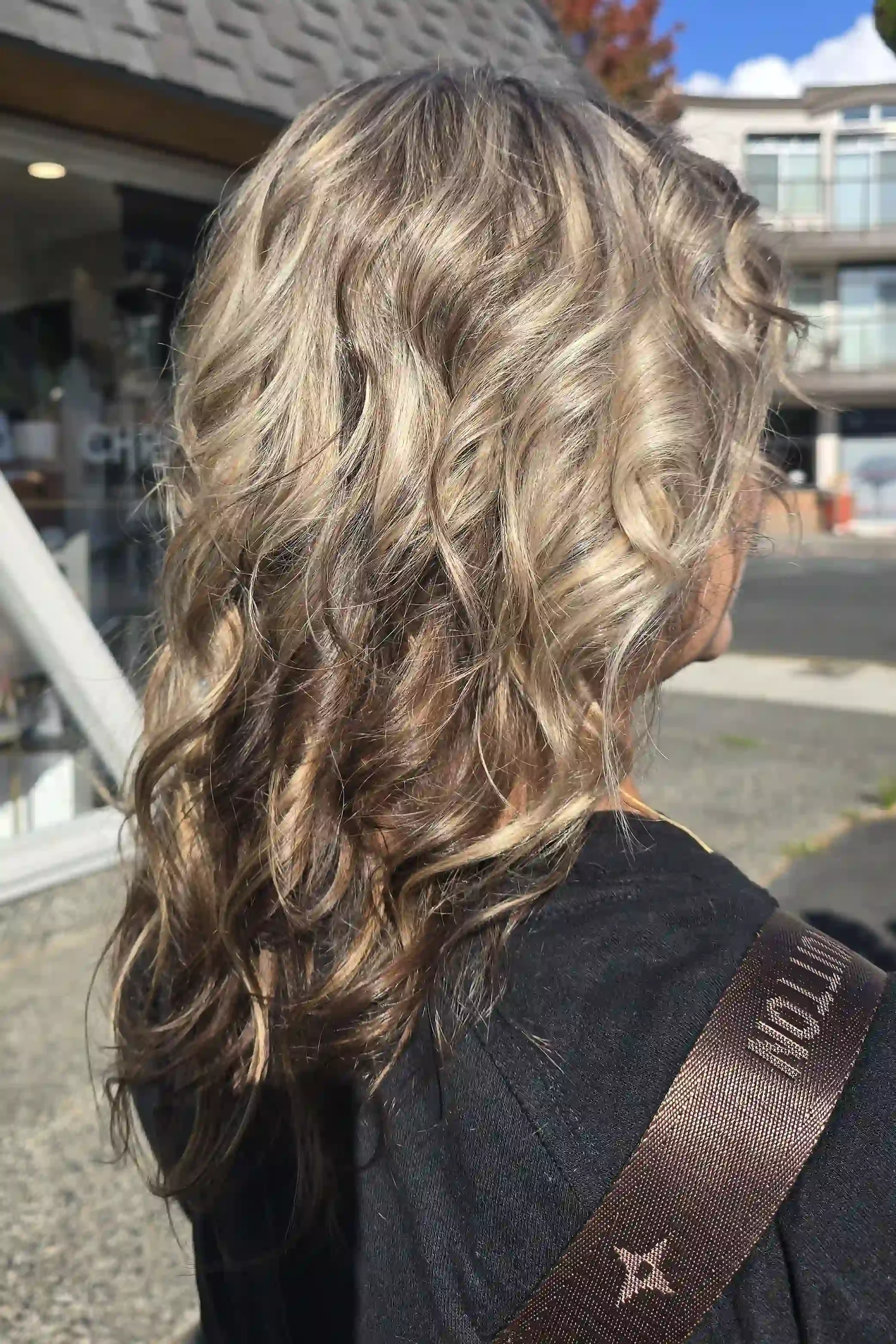 Dimensional ash blonde hair with bright champagne highlights woven through a deeper brunette base, creating beautiful contrast and depth. Long, cascading layers are styled in voluminous, defined curls that showcase the multi-tonal color and add movem