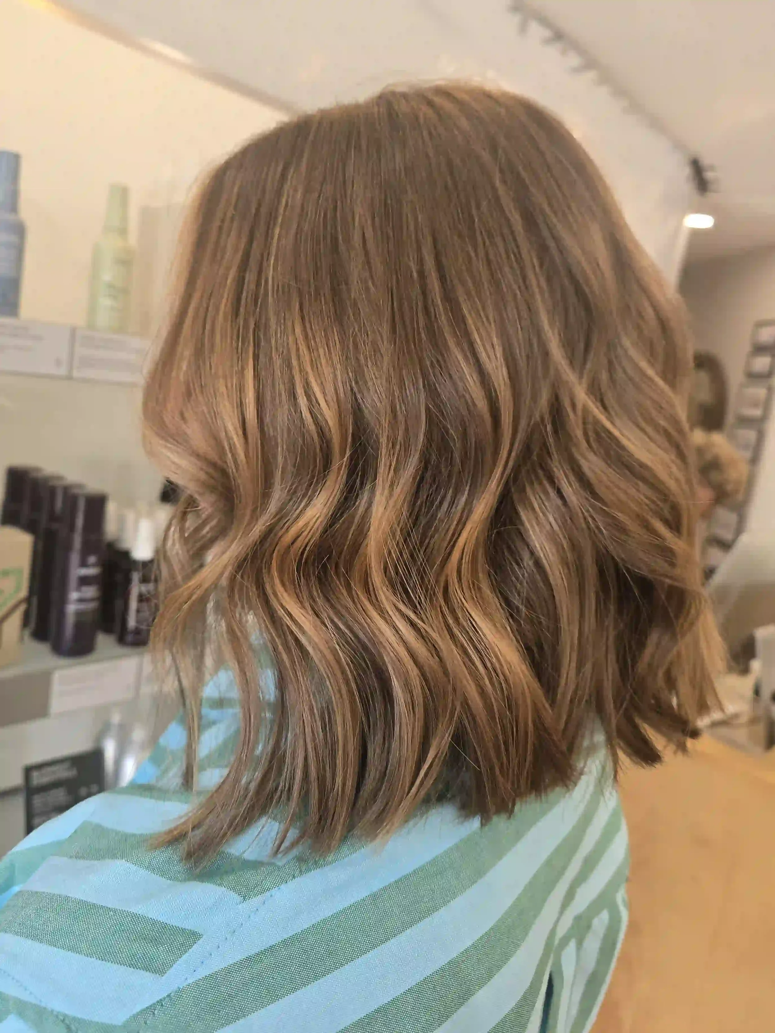 Warm brunette hair with soft caramel and copper-toned dimension, styled in loose, tousled waves. The textured, shoulder-length lob features subtle layers and softly blended ends for natural movement and a relaxed, effortless finish. Hair by Jhana at 