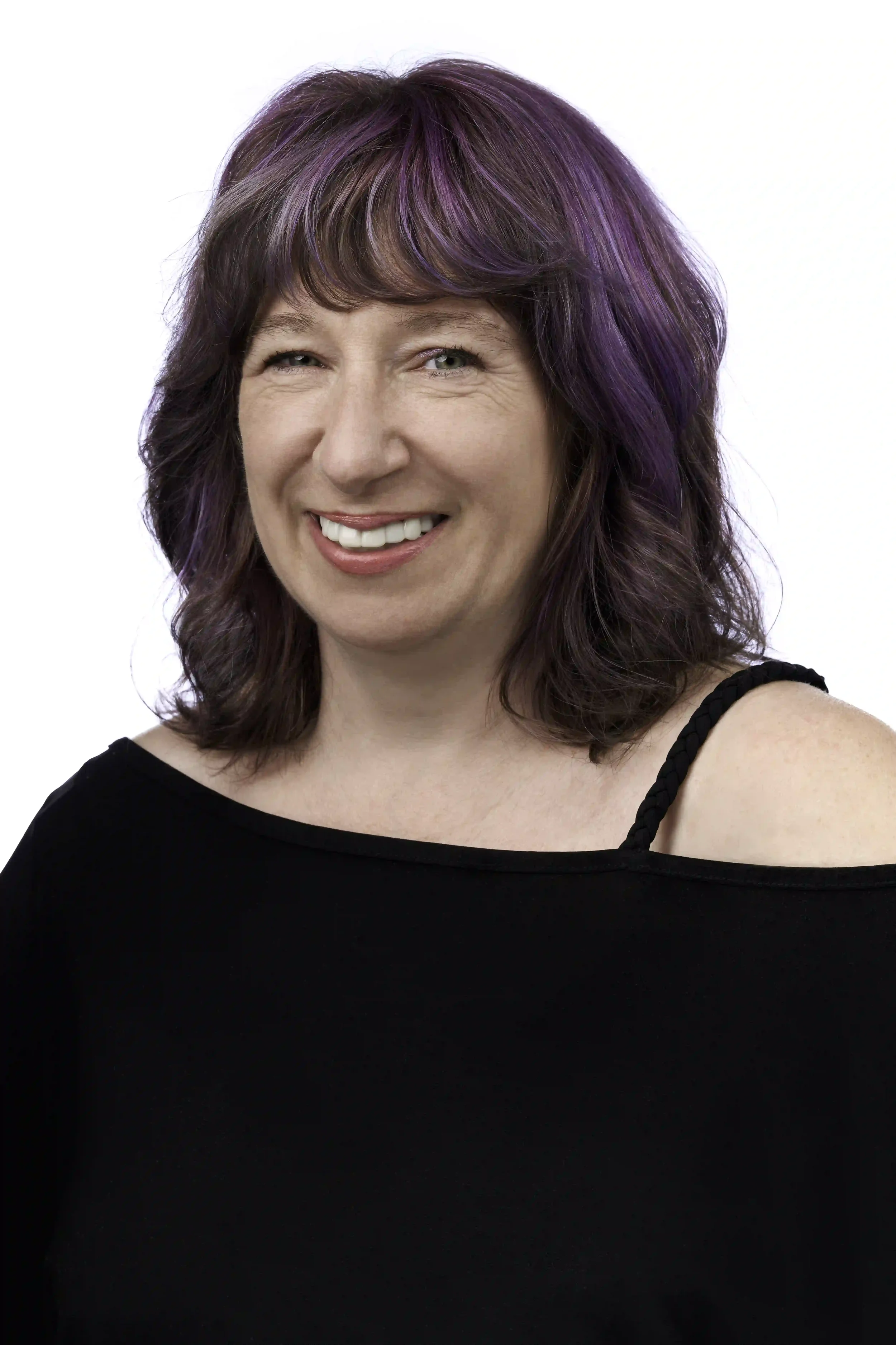 Smiling woman with wavy brown hair and purple highlights wearing an off-the-shoulder black top, photographed against a white background.