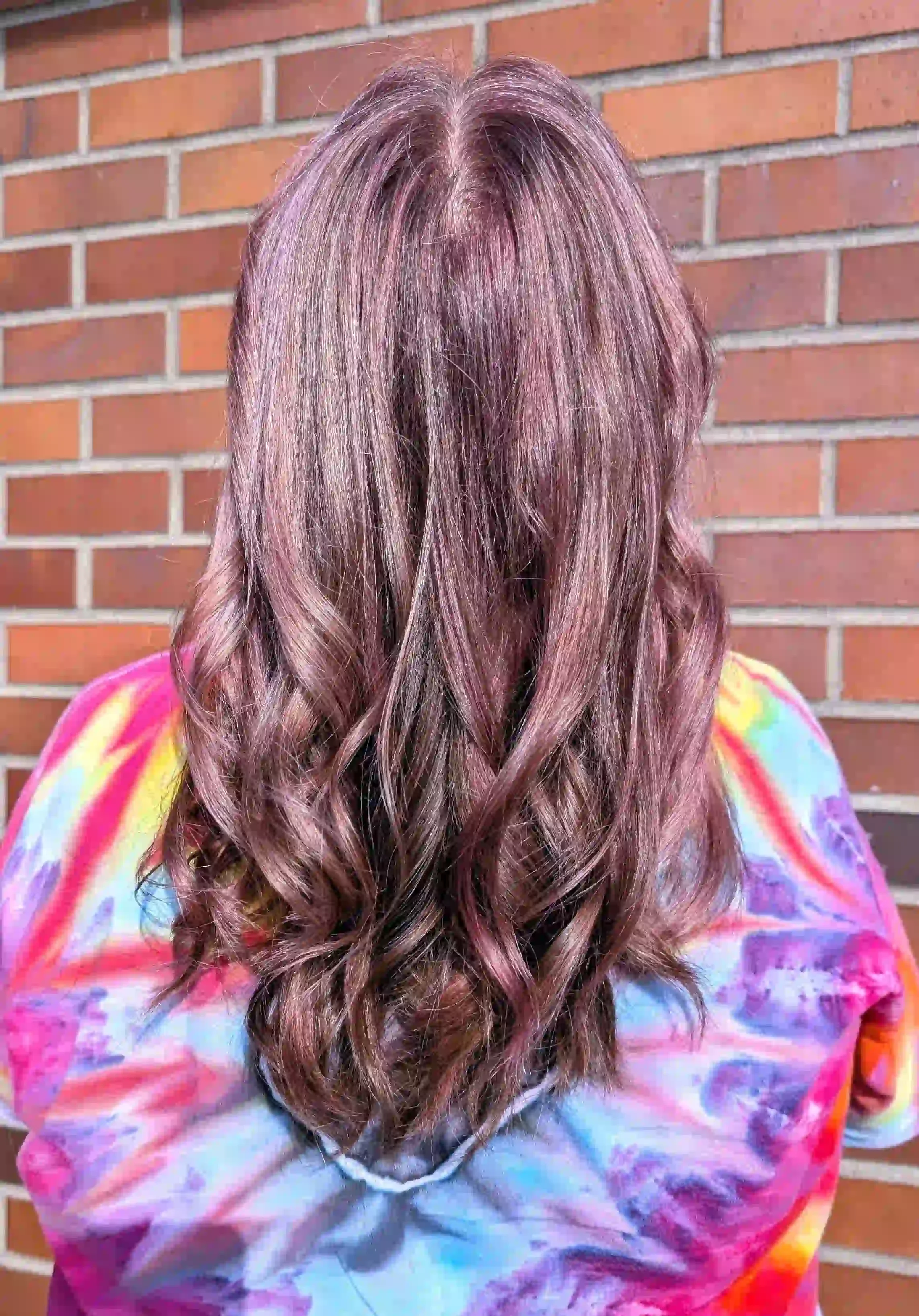 Soft dusty rose brunette balayage with mauve undertones and glossy dimensional waves. Long layered styling enhances the rosy shimmer and blended depth for a romantic, modern fashion color finish.