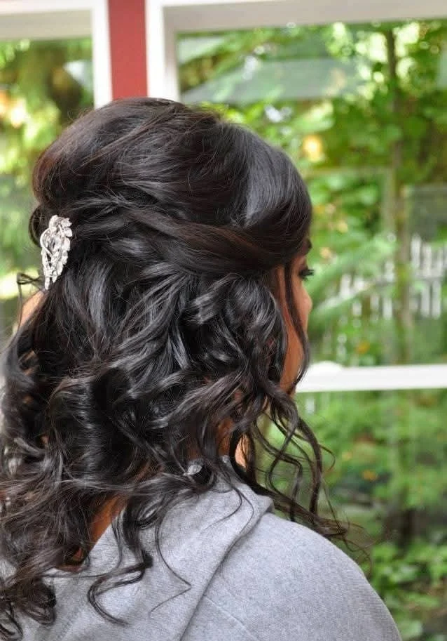 Elegant half-up bridal hairstyle featuring soft cascading curls, romantic twists, and a jeweled hair accessory for a timeless wedding-day look. This classic bridal style combines polished volume, graceful movement, and intricate detailing to create a