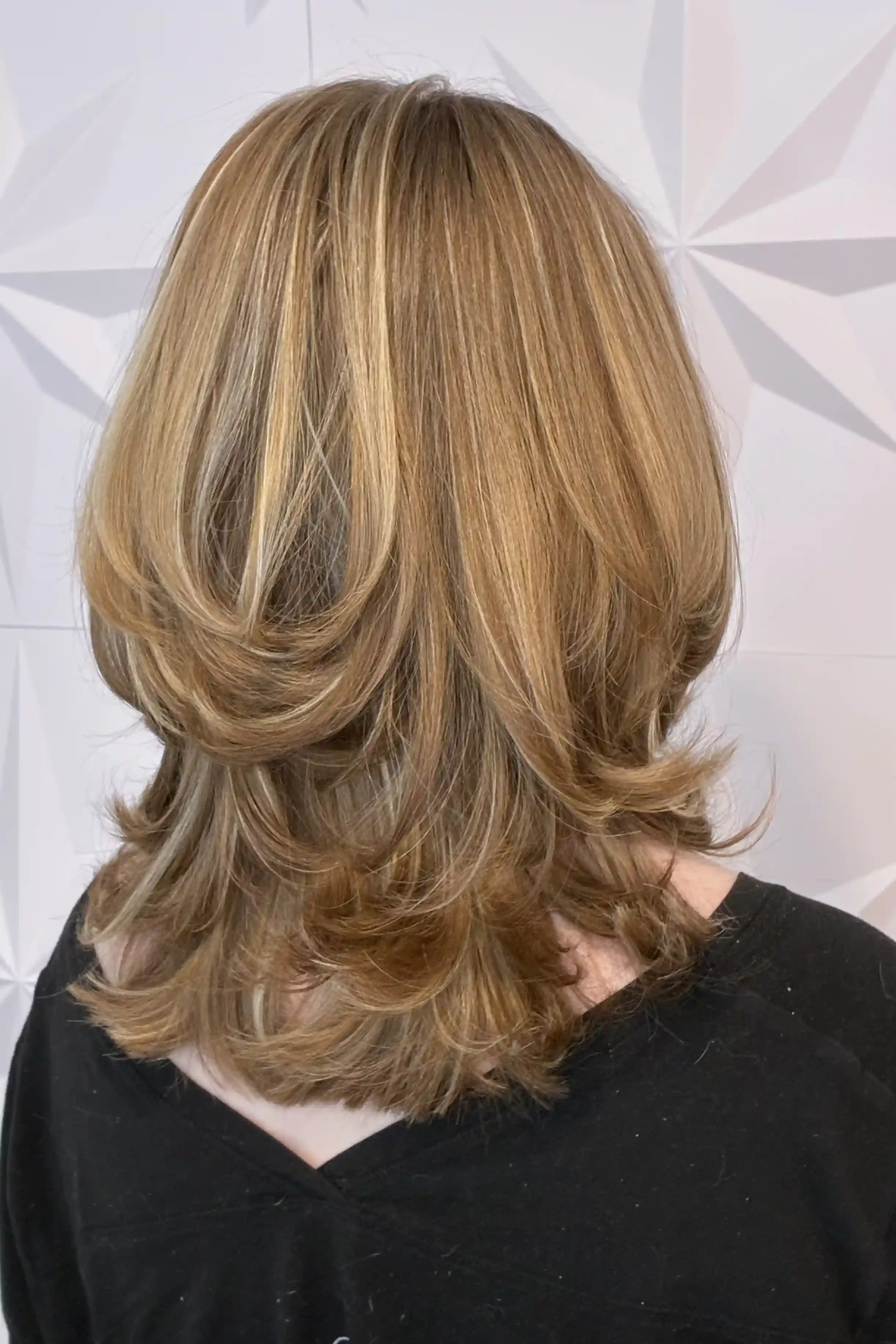 Golden bronde hair with soft blonde and caramel dimension, cut into long cascading layers and styled with a bouncy, rounded blowout. The layered shape creates fullness, movement, and a polished finish with softly flipped ends. Hair color, haircut, an