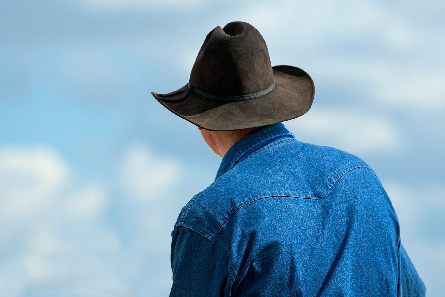 A photograph of a western cowboy and his black cowboy hat shot from behind on a Montana ranch.&nbsp;→ Buy a Print
