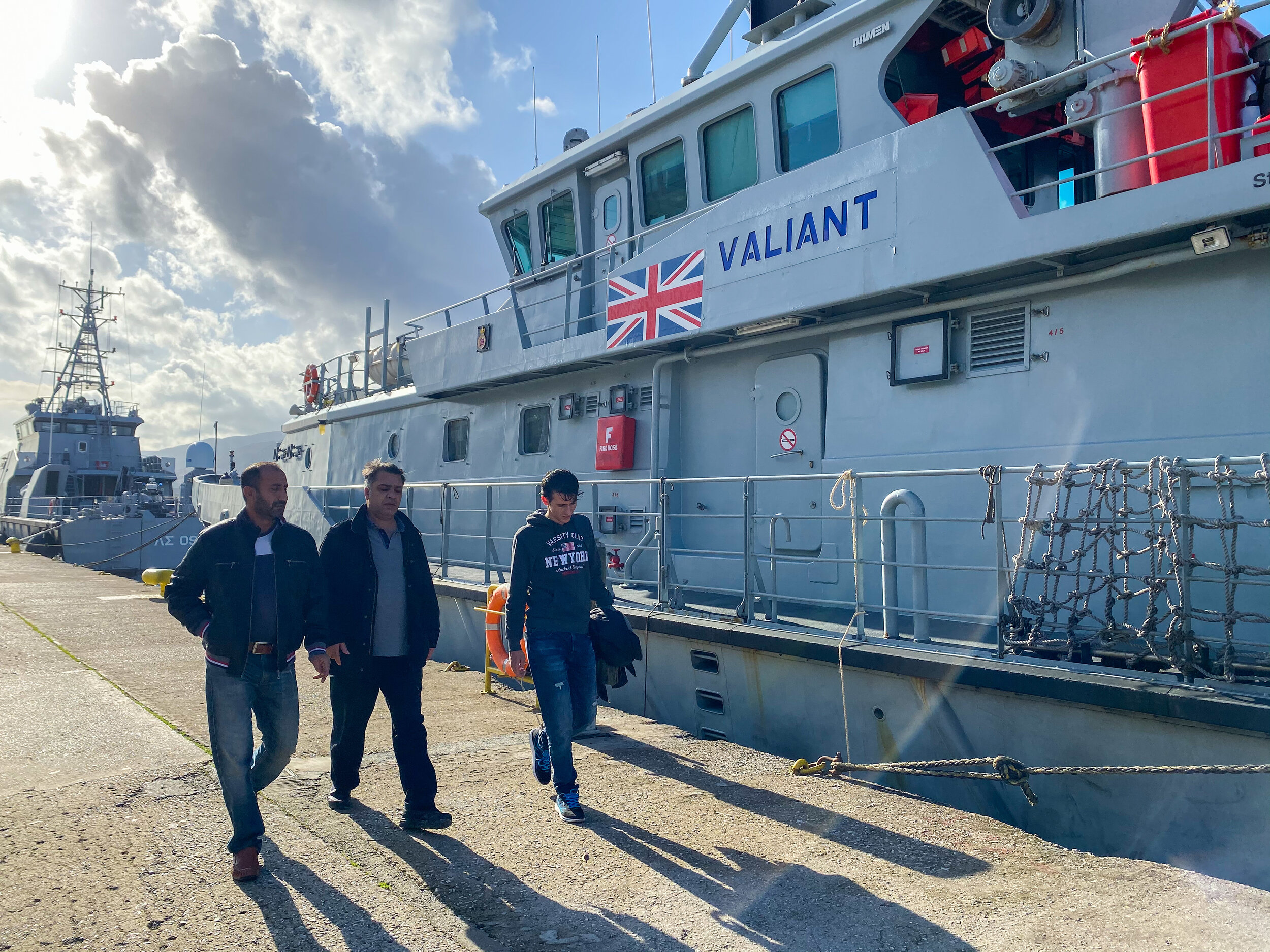 Asylum seekers walking past a British border patrol boat in the port of Mytilini on Lesvos island. Photo Credit: Devon Cone.