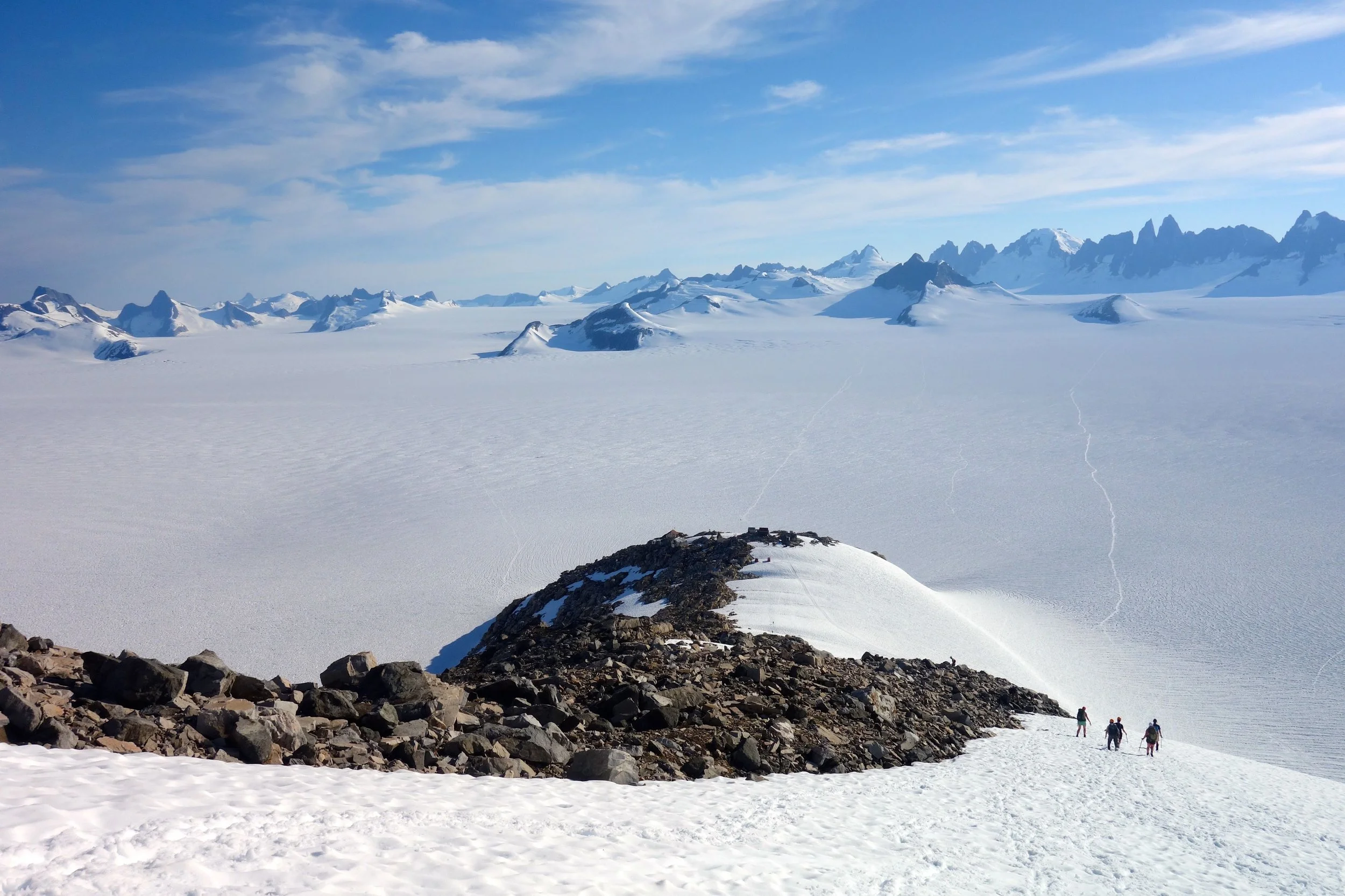 The hike down to camp for dinner. At the top of the rocky hilltop in the fore ground you can pick out the buildings of our largest camp. Beyond camp, Taku Glacier flows from right to left in front of the distant Taku Towers. Photo credit: Kenzie McAdams.