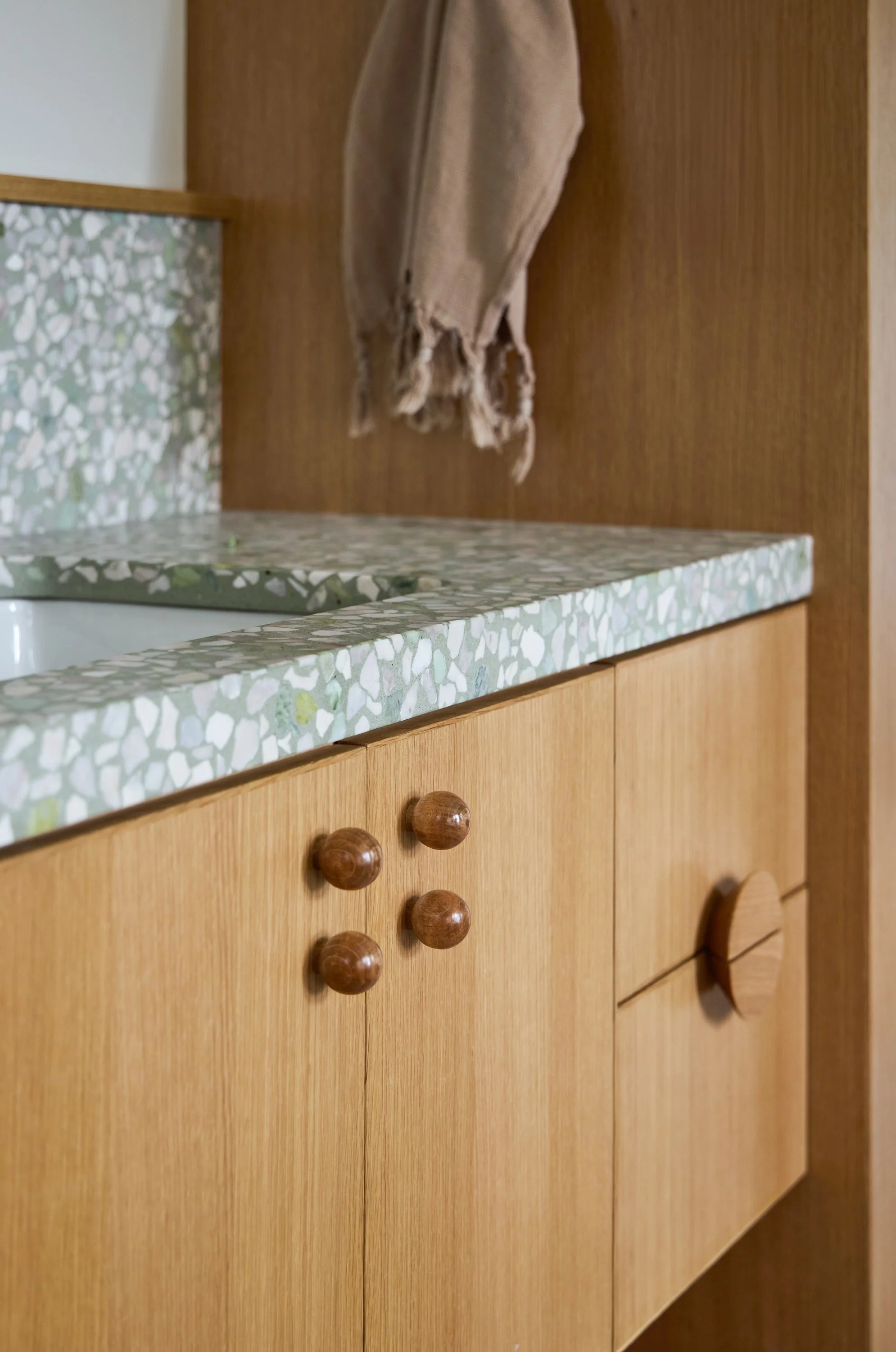 A close-up of a wooden kitchen cabinet with round wooden knobs, a textured mosaic countertop, and a beige towel hanging on a wooden panel behind it.