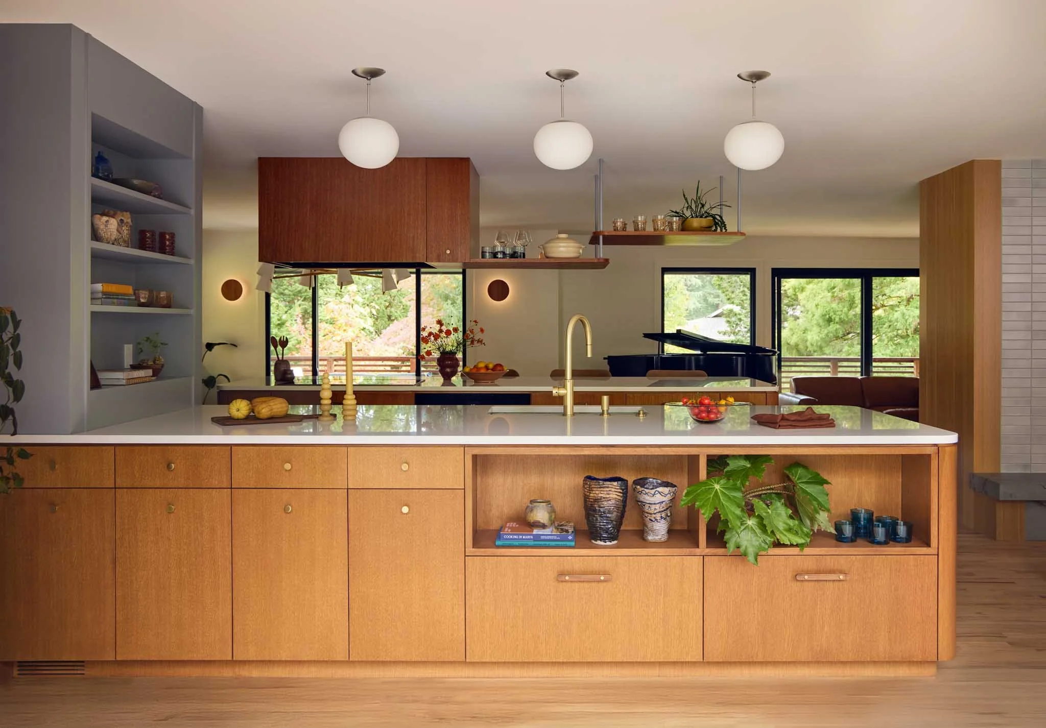 Modern kitchen with wooden cabinets and a white island counter, decorated with pumpkins, candles, and a bowl of apples, overlooking large windows with views of green trees outside.