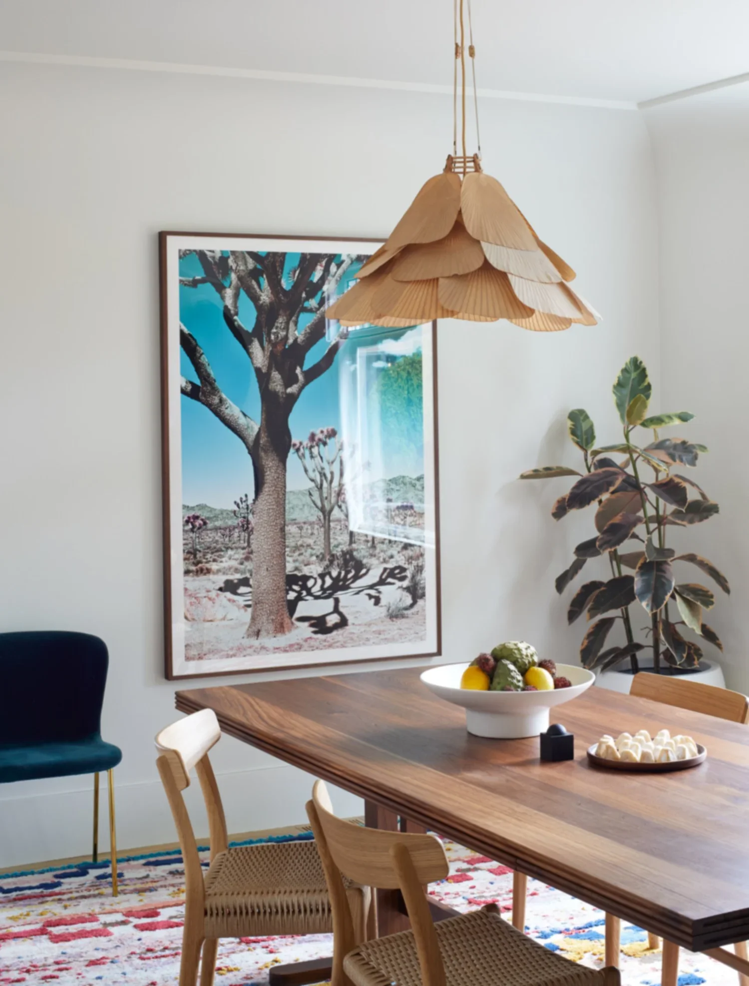 A dining room with a large wooden table, a hanging leaf-shaped light fixture, a bowl of fruit, a small black box, and a plate with meringues. A large framed desert landscape with a tree is on the wall, and a potted plant is in the corner.
