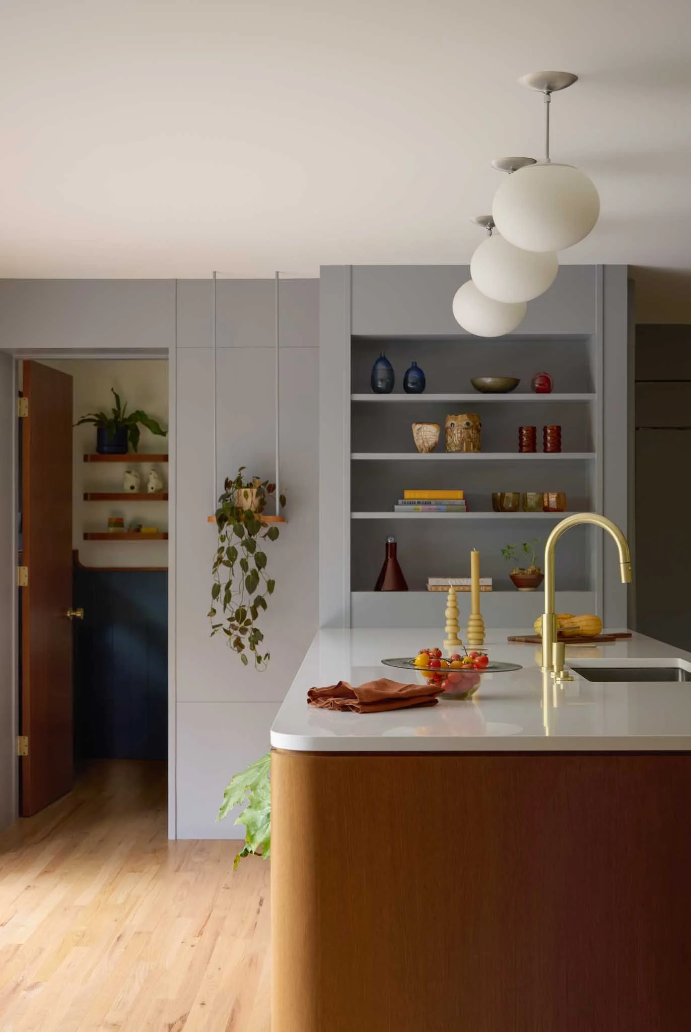 Modern kitchen with a white island, gold faucet, hanging white globe pendant lights, open shelving with decorative vases, bowls, and books, and wooden flooring.