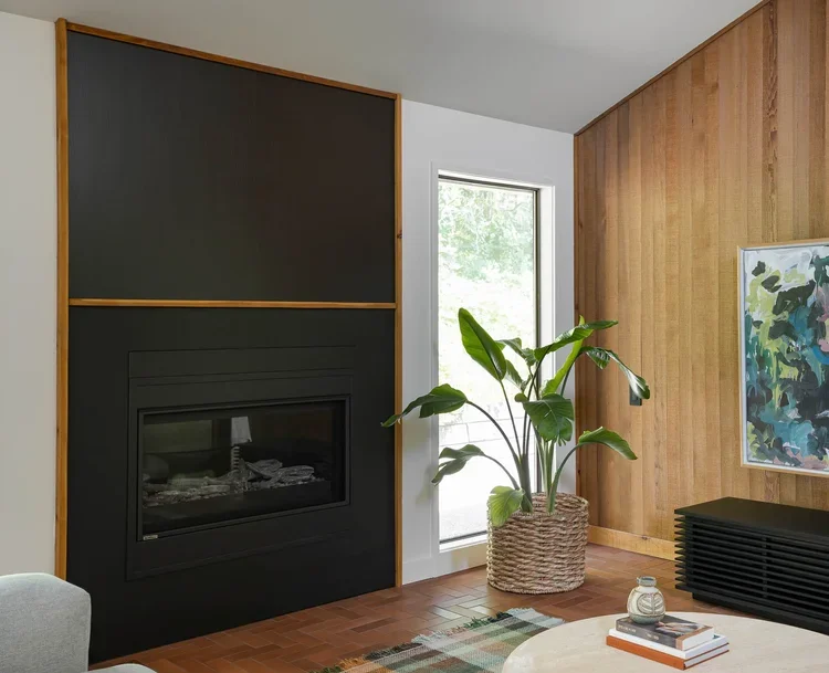 Interior of a living room featuring a modern black fireplace with a wood frame, a large window letting in natural light, a potted green plant, a colorful abstract painting on a wood-paneled wall, and a small black cabinet.
