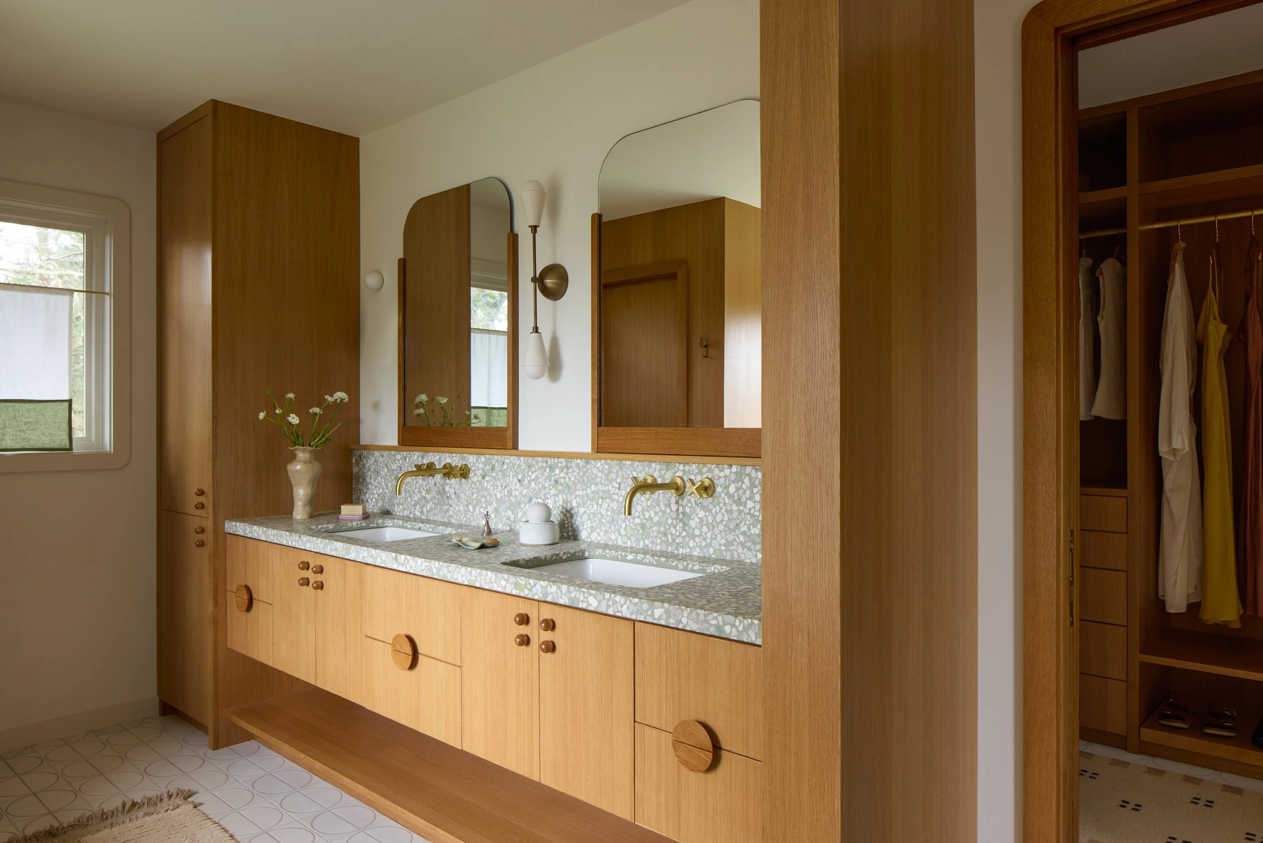 Bathroom with wooden cabinetry, a double sink with a terrazzo countertop, gold fixtures, a large mirror, a vase with flowers, and a walk-in closet with hangers and clothes.
