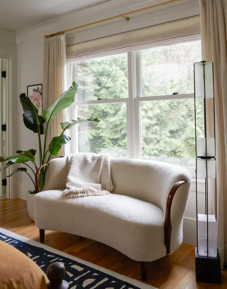 A cozy living room corner featuring a cream-colored vintage loveseat, a tall potted plant with large green leaves, a window with white curtains, and a modern floor lamp with multiple tiers of white rectangular shades.