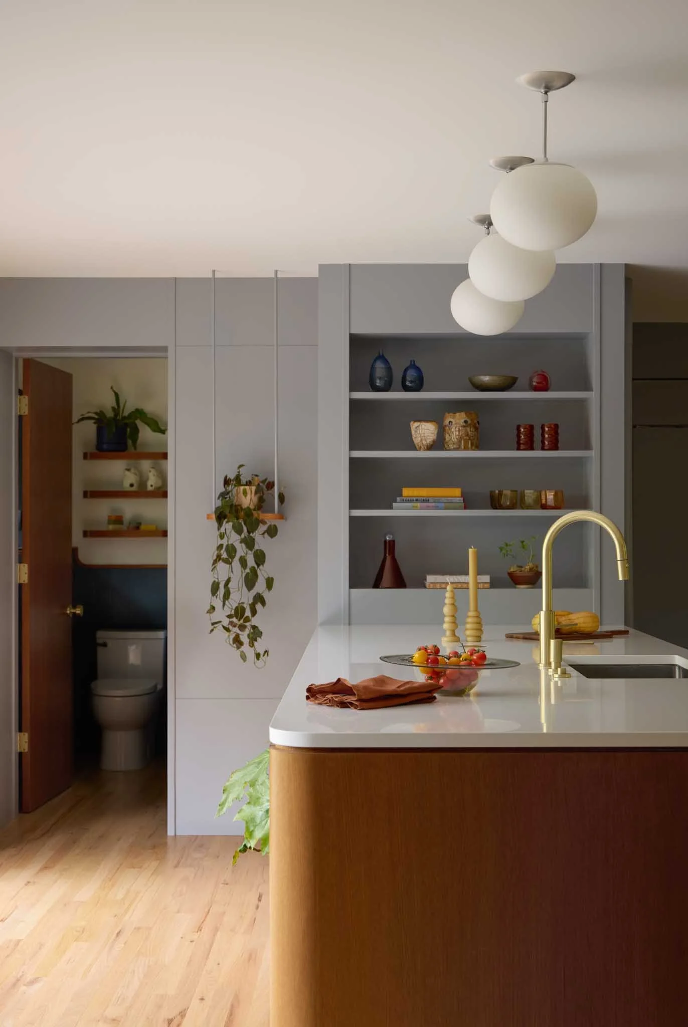 Modern kitchen with a white island, gold faucet, open shelving with decorative items, and a view into a small bathroom with a toilet. Light wood flooring, plants, and minimal decor.