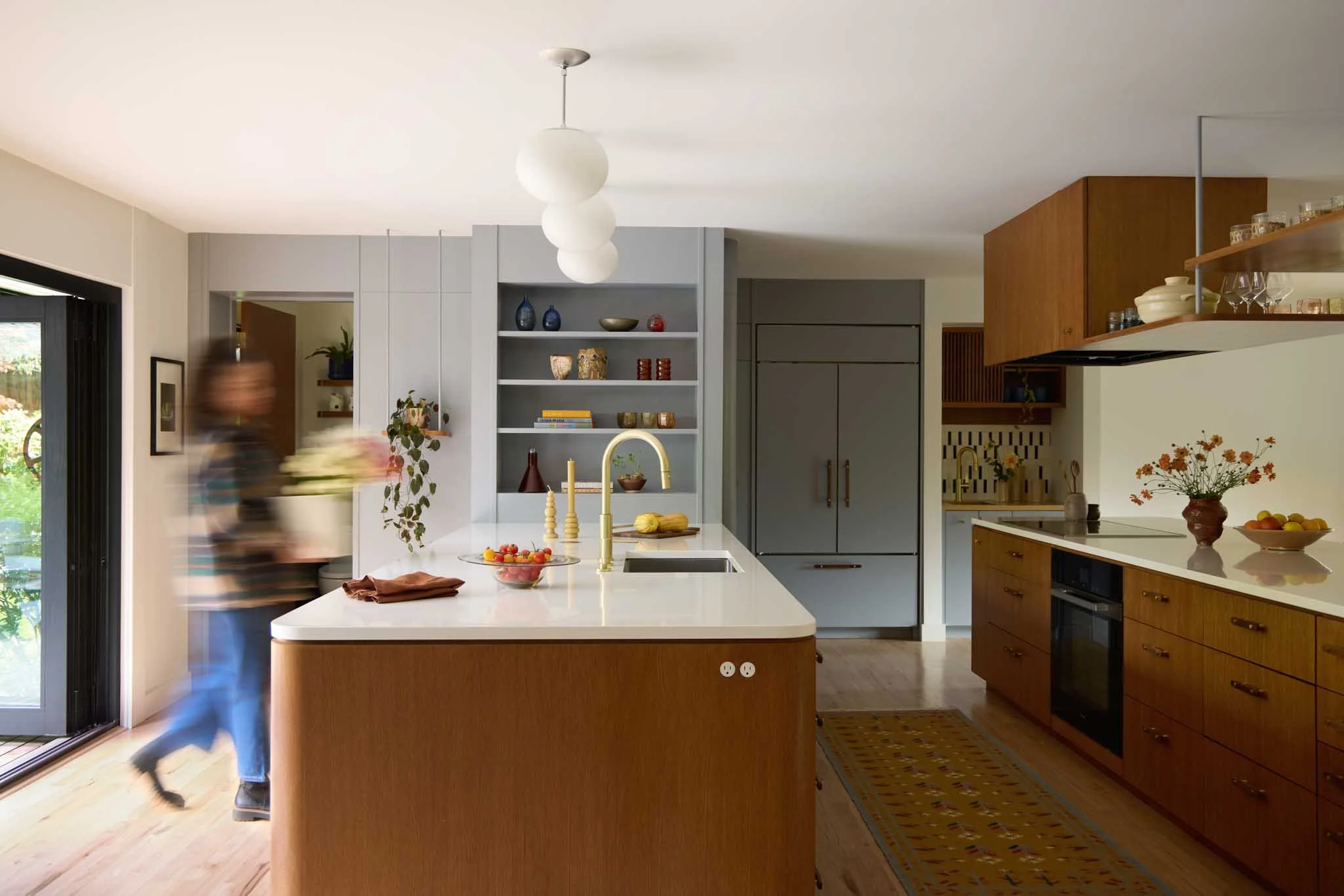 A modern kitchen with a white island countertop, gold faucet, open shelves with decorative items, pantry, and wooden cabinetry. A person is blurred in motion near the sliding glass door.