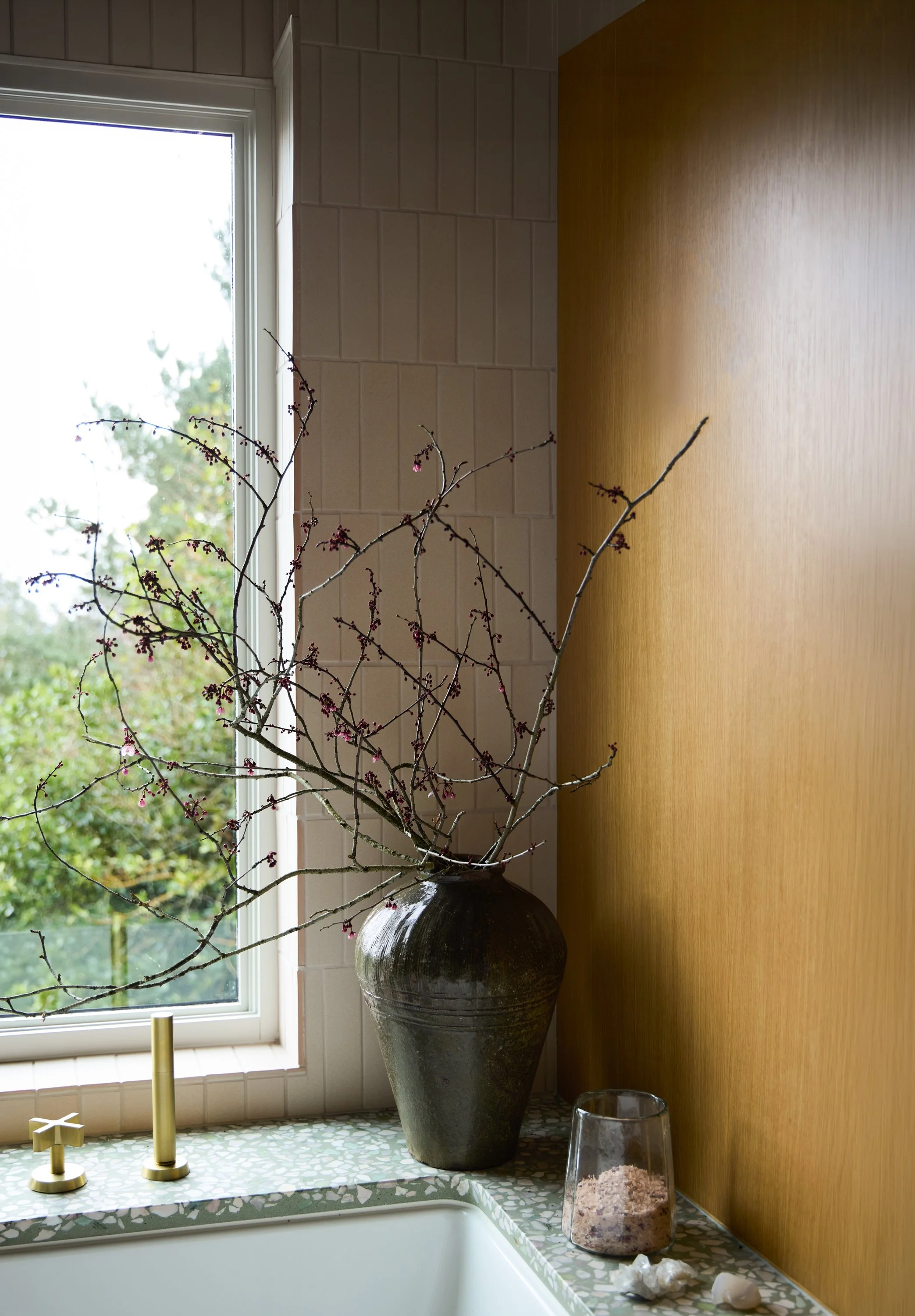 A kitchen window with a white frame, overlooking green trees outside. On the window sill, there's a large dark ceramic vase containing bare branches with small pink flowers. The counter beneath the window has a green and white mosaic tile pattern, with items including a gold candle holder with a single candle, a glass container filled with pink salt, and some small white decorative rocks.