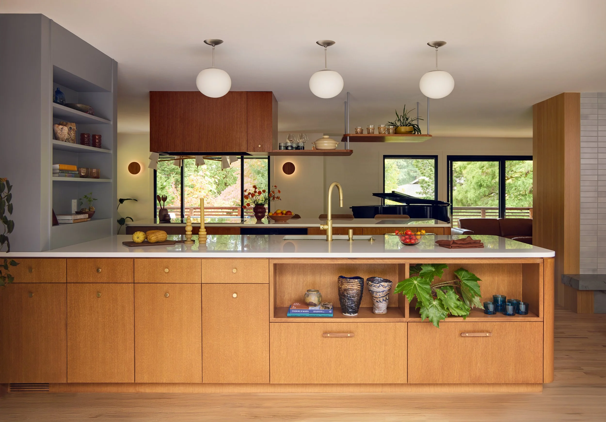 Modern kitchen with wood cabinetry, large island, white countertop, gold faucet, and greenery. Open shelves with decor and dishes, large windows with a view of trees, and hanging globe lights.