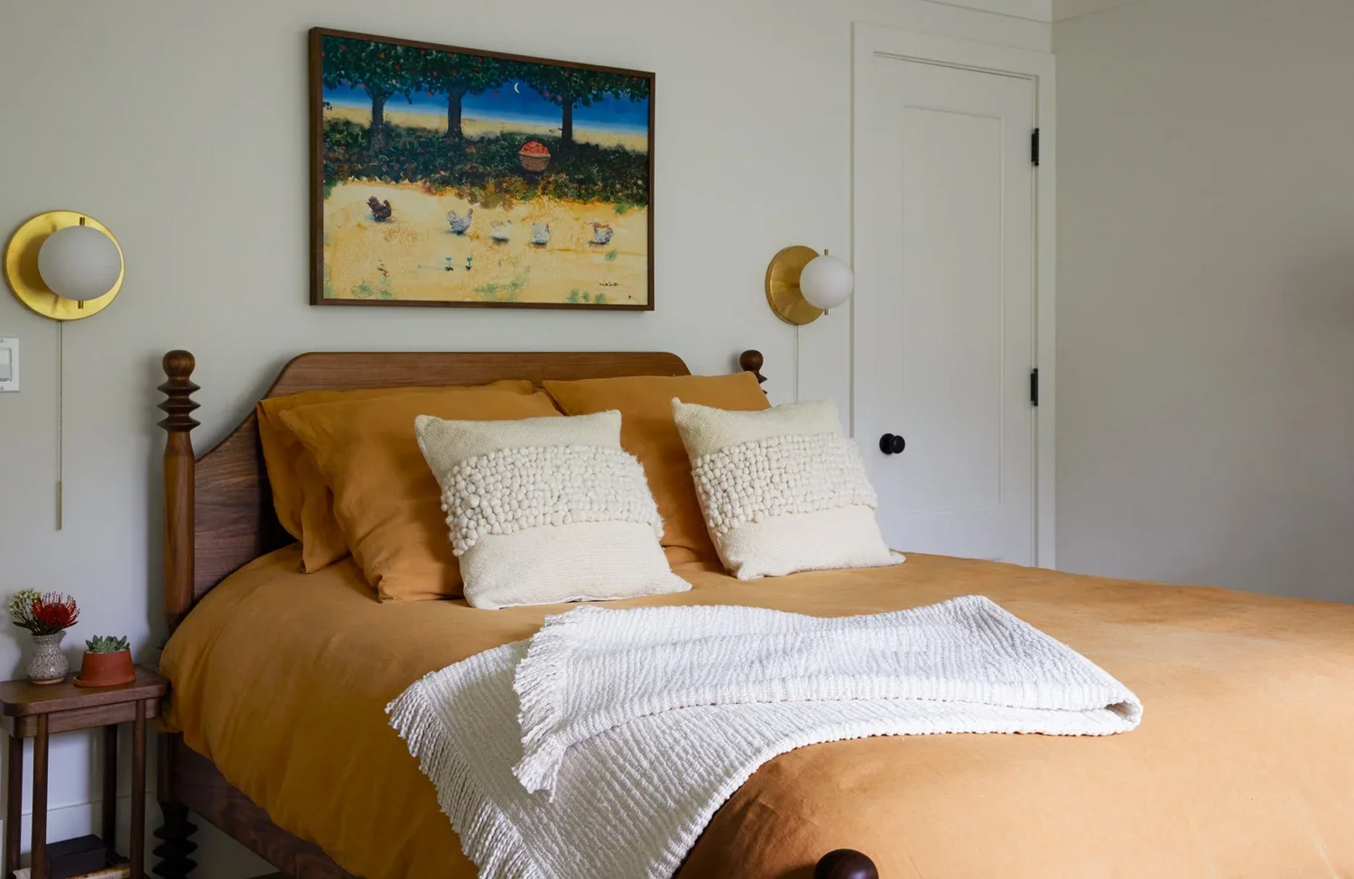 A neatly made bed with brown bedspread and two beige decorative pillows, a white throw blanket at the foot, flanked by two wall-mounted lights and a small wooden nightstand with plants, with a colorful landscape painting above.