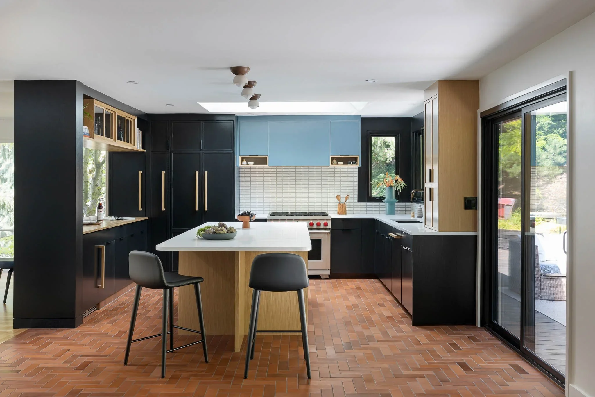 Modern kitchen with black and light wood cabinetry, white countertops, brick-patterned flooring, a kitchen island with two black chairs, a blue upper cabinet, white backsplash tiles, and large windows and sliding glass door bringing in natural light.