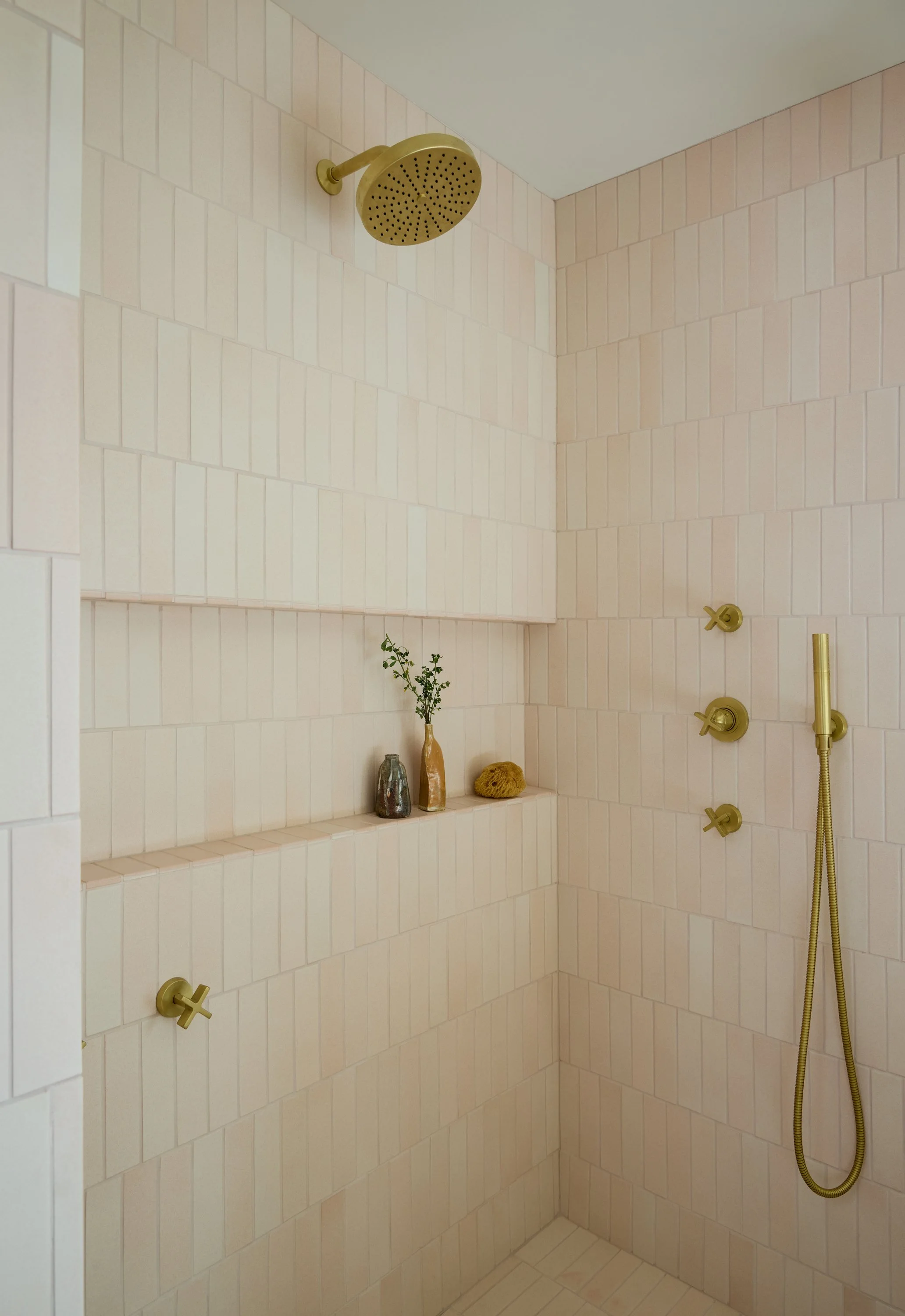 Shower area with beige tiled walls, gold fixtures, a showerhead, a handheld shower, and a small shelf with vases and decorative objects.