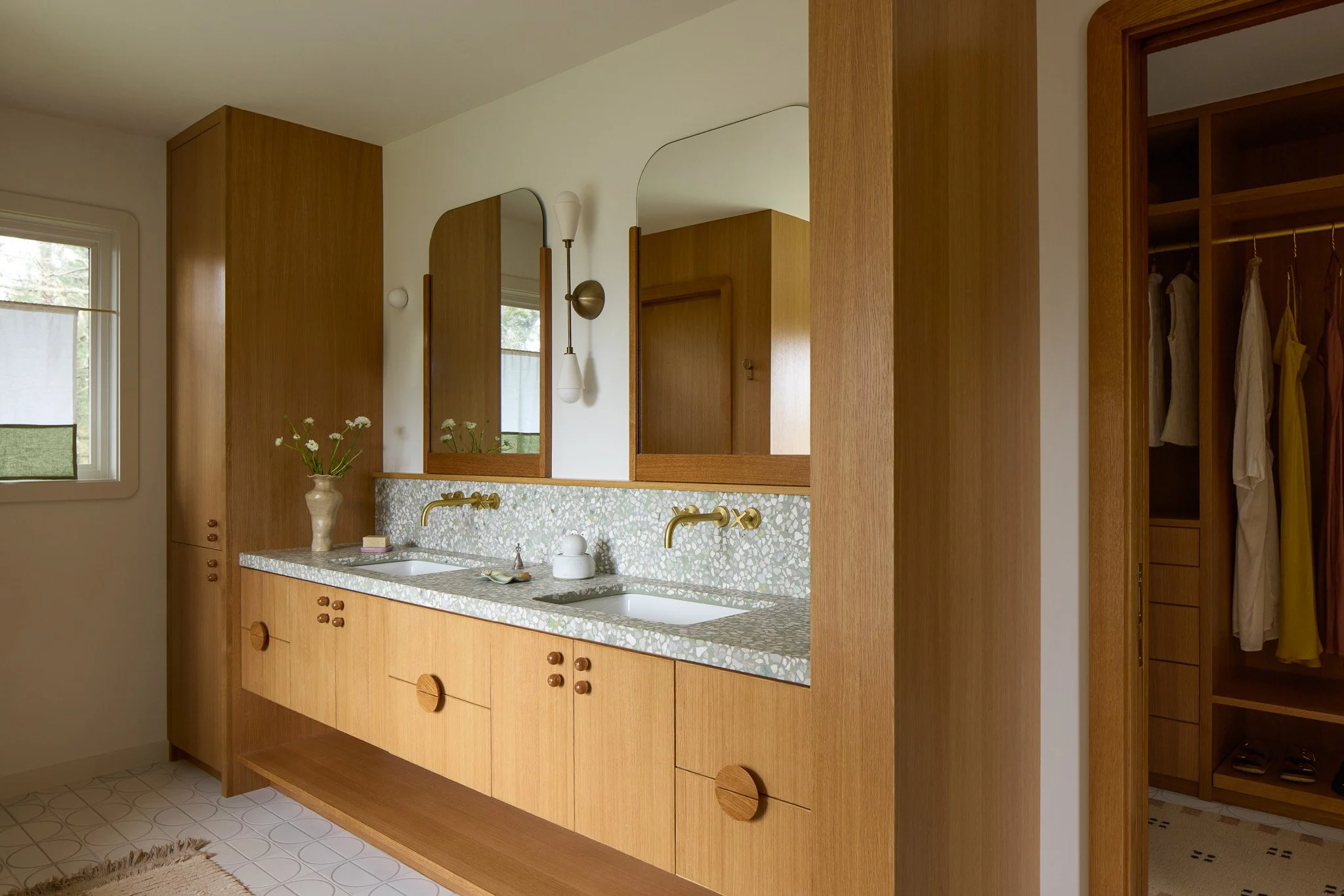 A mid-century modern bathroom with honey-colored wooden cabinetry, terrazzo countertops, and brass fixtures. Decor includes a white vase with flowers, a soap dish, and natural light from a nearby window.