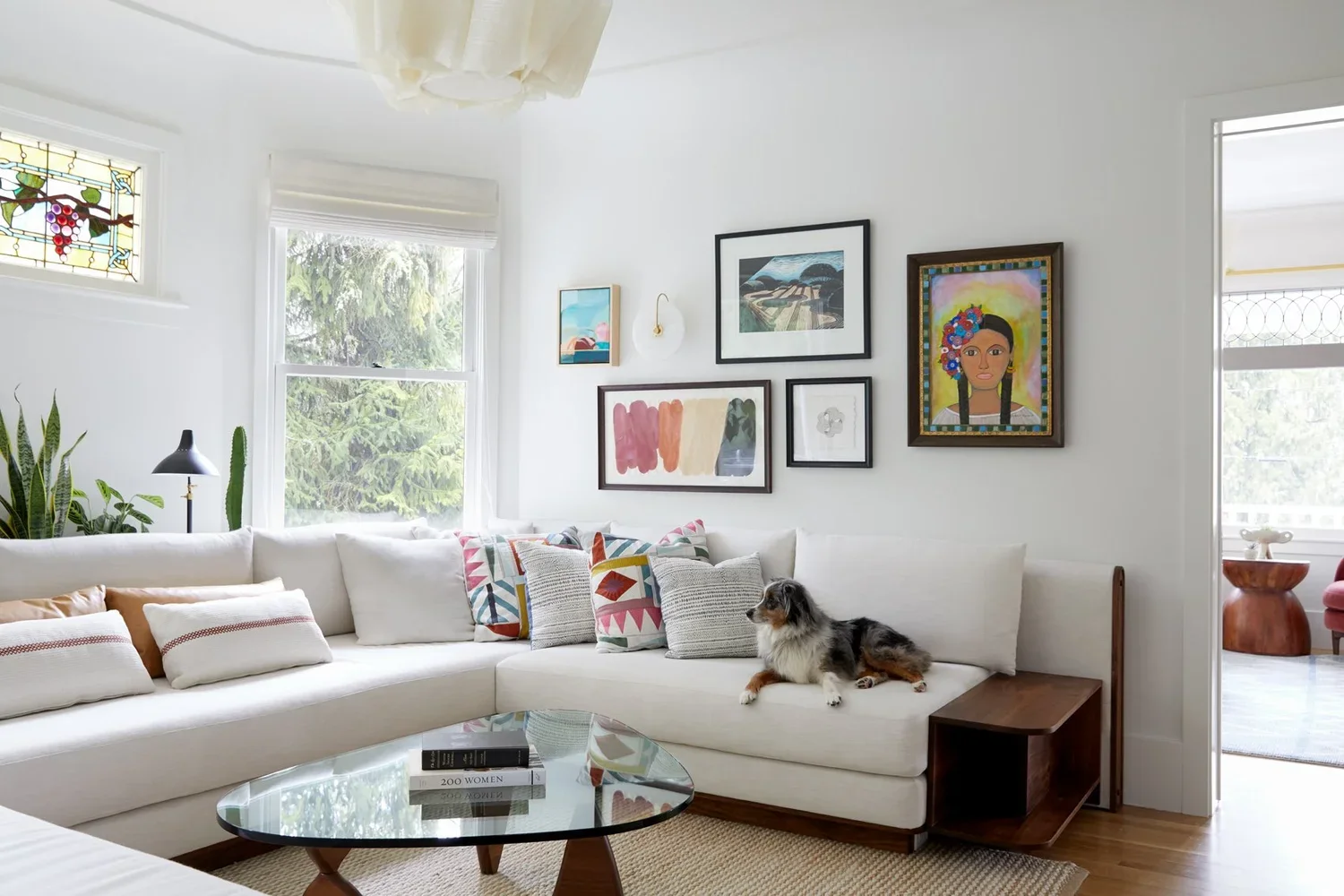 Bright living room with white sectional sofa, colorful pillows, framed art on the wall, and a dog resting on the sofa. There are windows with greenery outside and a glass coffee table with books.