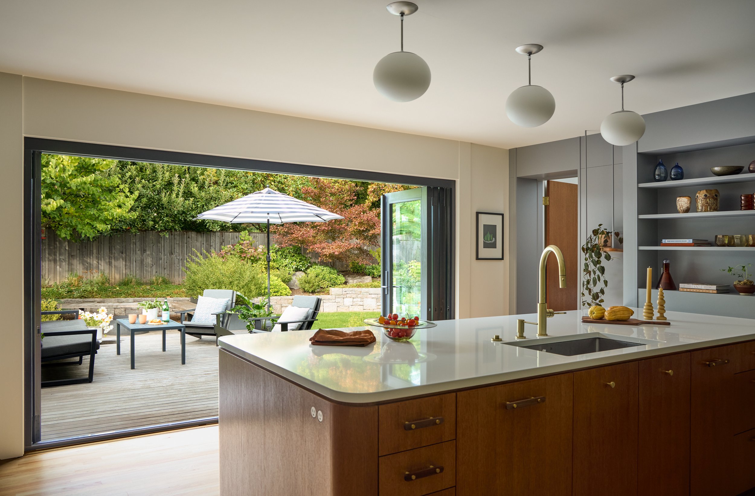 View of a modern kitchen with an open sliding glass door leading to a backyard patio with outdoor furniture, umbrellas, and lush greenery.