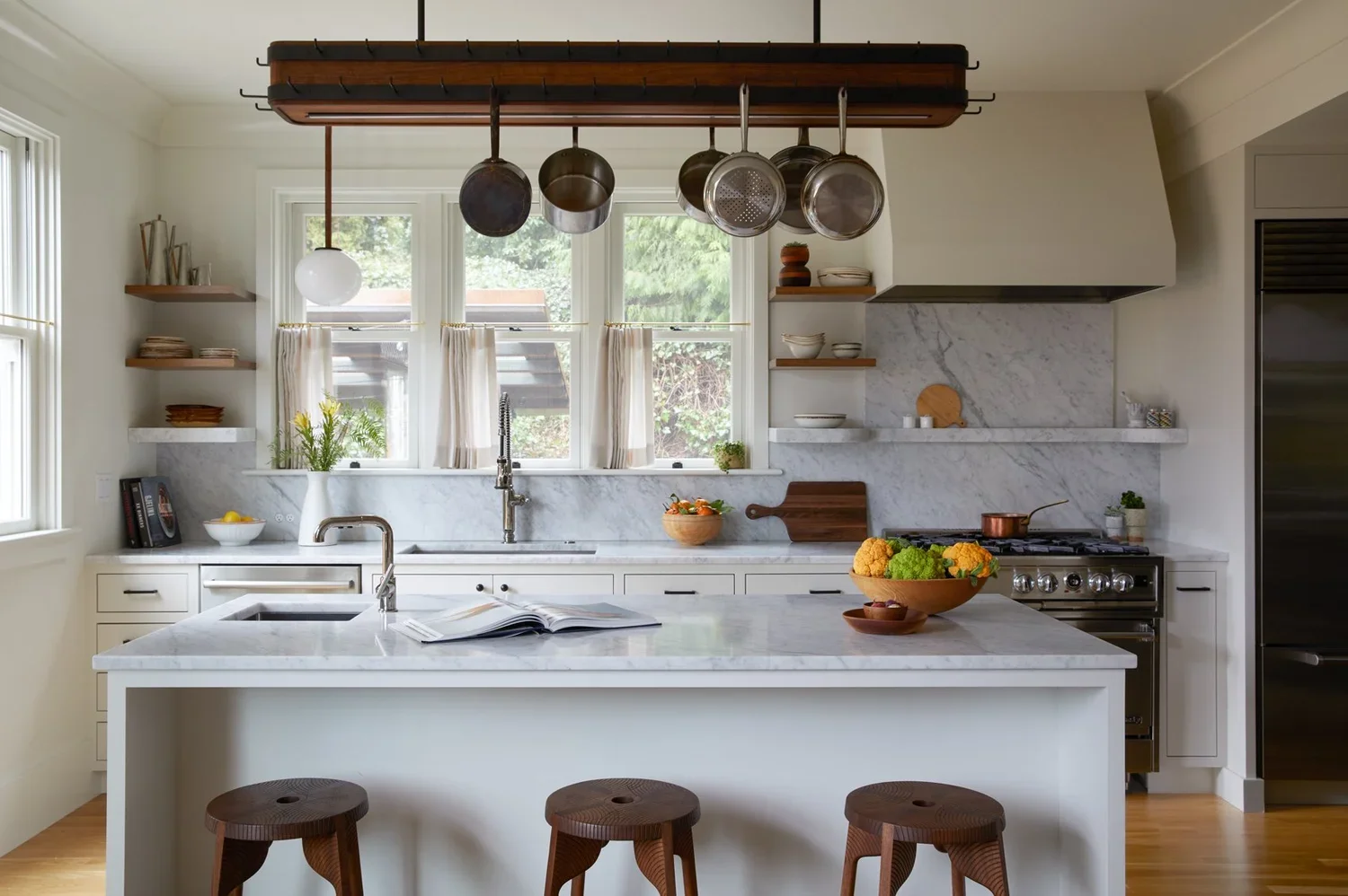 A bright kitchen with a white marble island, open shelves, and a window above the sink. Various pots and bowls are on shelves and counters, with a bowl of vegetables on the island and a cookbook open. Three wooden stools are at the island, and hanging pots and pans are above.