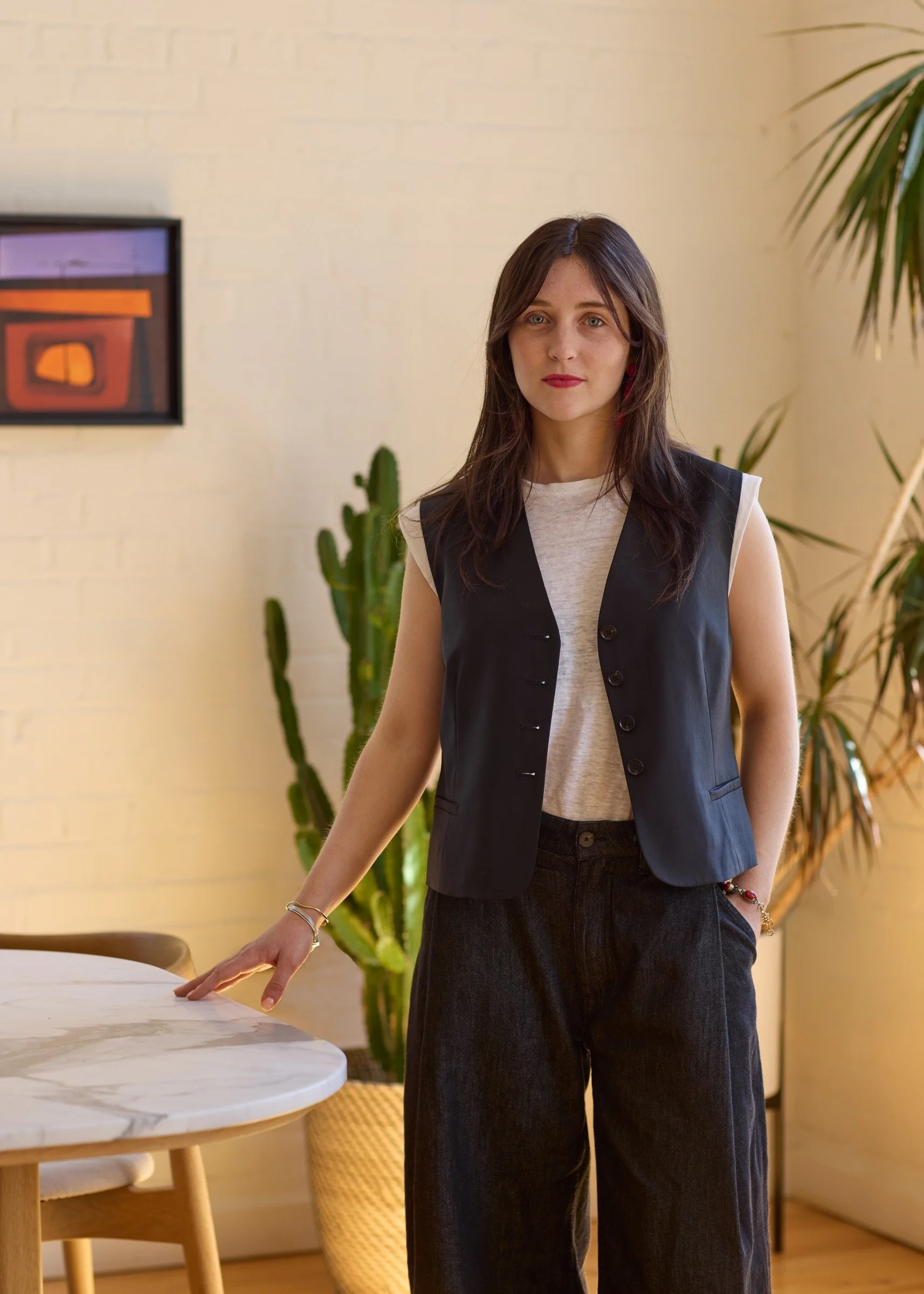 A woman with long brown hair, wearing a light beige shirt and a black vest, standing indoors next to a table, with her hand resting on the table, in front of a beige wall with a framed abstract art piece and large green plants in the background.